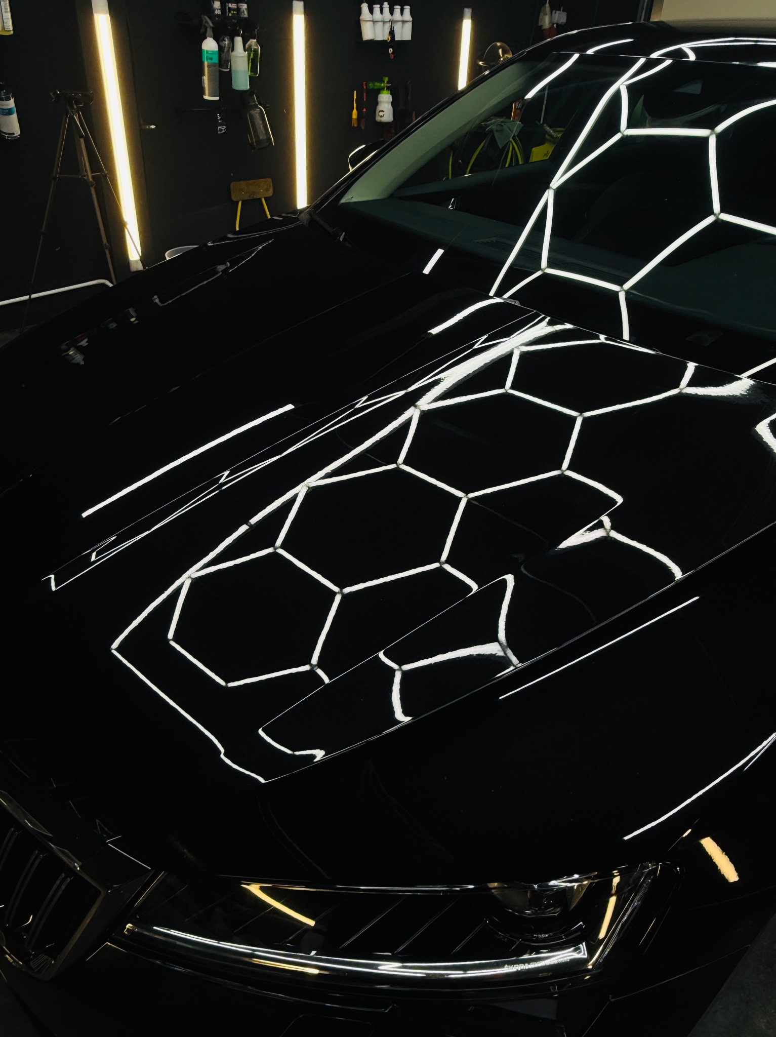 Błyszczący, czarny samochód po detailingu, z odbiciami lamp w kształcie heksagonów na masce. Widoczne reflektory i fragment grilla. W tle warsztat z narzędziami.