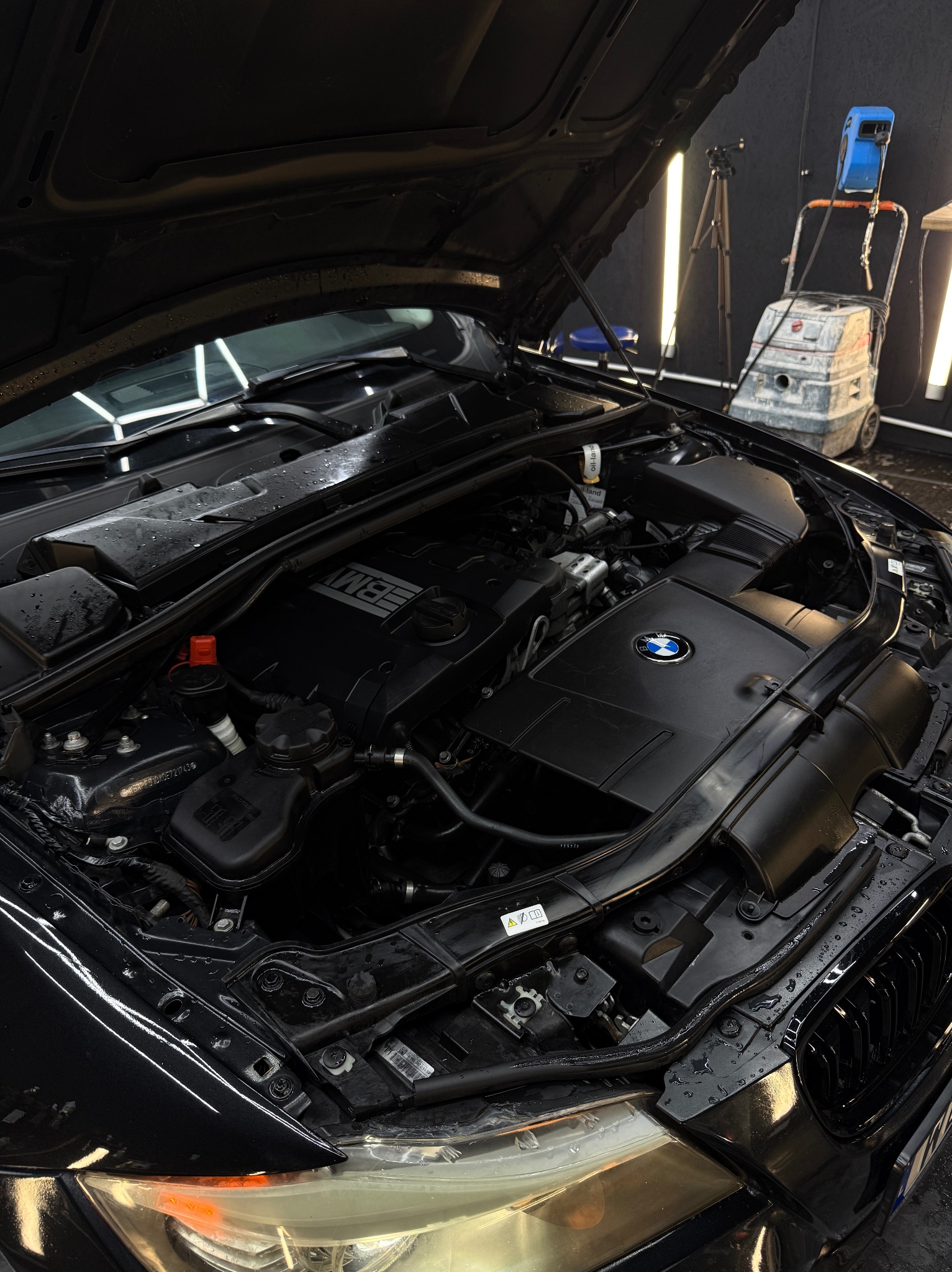 Otwarta maska czarnego BMW, widoczny silnik po czyszczeniu, krople wody na elementach. W tle sprzęt do czyszczenia.