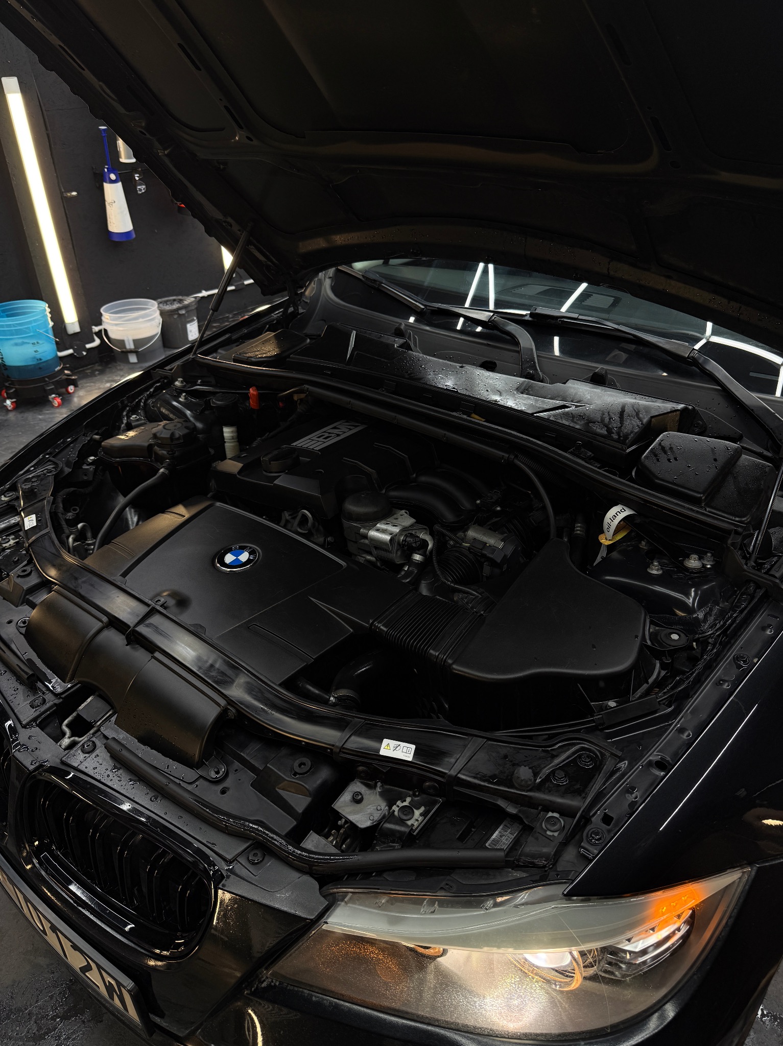 Czarny samochód BMW z otwartą maską, widoczny silnik po czyszczeniu. Krople wody na elementach, oświetlenie warsztatowe w tle. Detailing silnika.