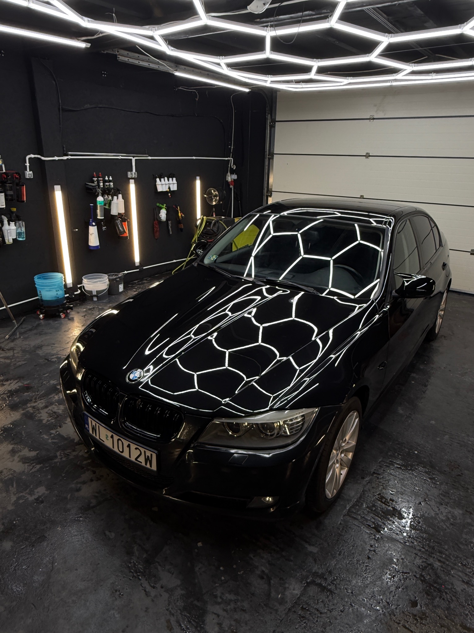 Błyszczący, czarny samochód BMW po detailingu w myjni. Na lakierze odbija się heksagonalne oświetlenie. Widoczne narzędzia i chemia na ścianie.