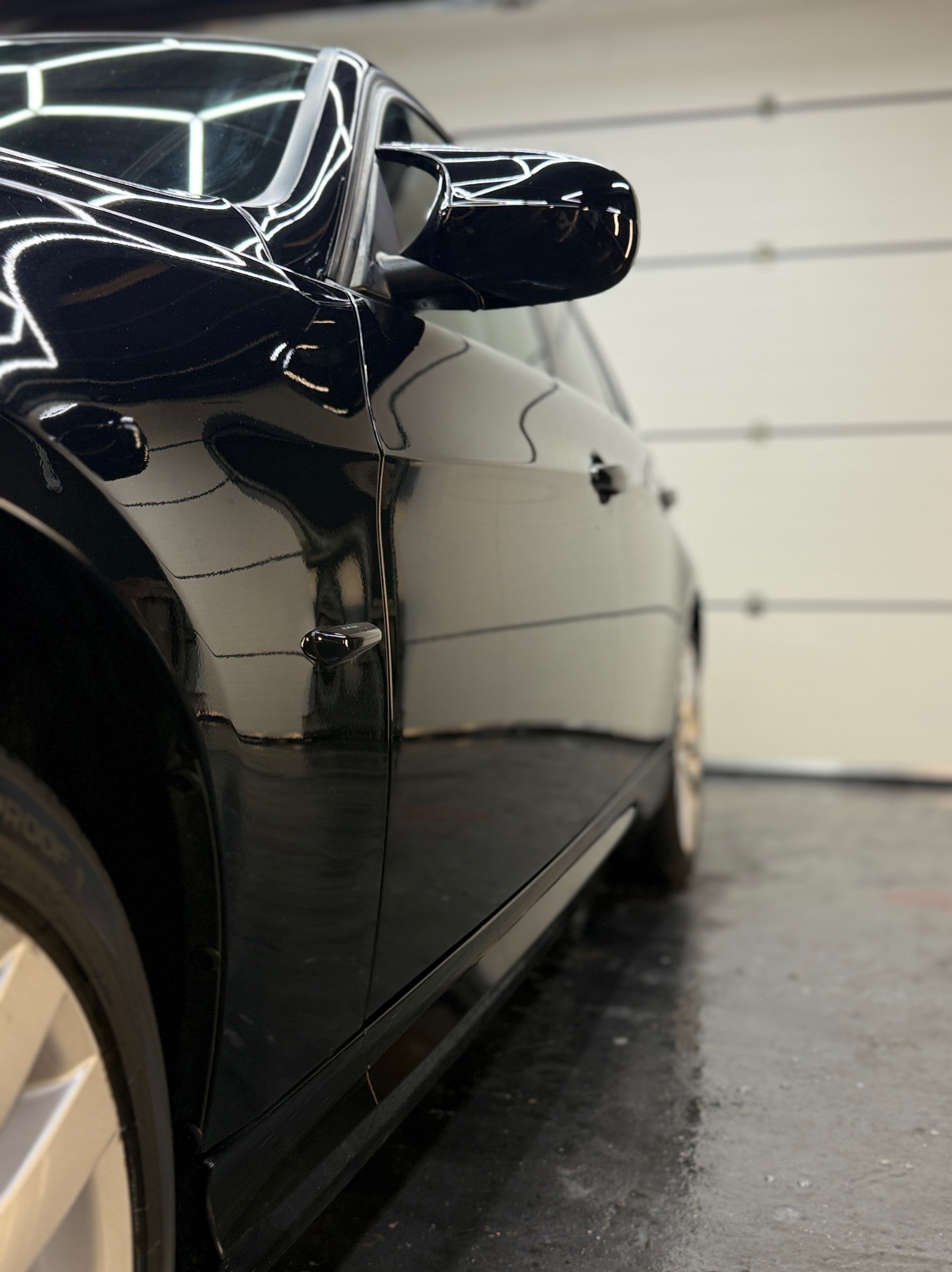 Błyszczący, czarny samochód po myciu, widok z boku, z refleksami światła na lakierze. Widoczna gładka powierzchnia i czyste felgi. Auto w garażu z białą bramą.