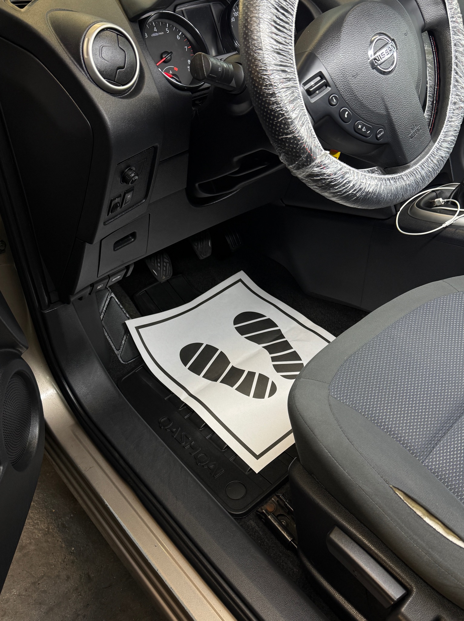 Wnętrze Nissana Qashqai z zabezpieczonym folią kołem kierownicy i papierową matą pod nogami kierowcy. Widoczny fragment fotela i deski rozdzielczej.