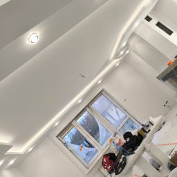 Łukasz Książek - Białe, nowoczesne wnętrze po malowaniu w Długosiodle. Sufit z oświetleniem punktowym i listwami. Okno zabezpieczone folią malarską. Widoczne narzędzia i materiały malarskie.