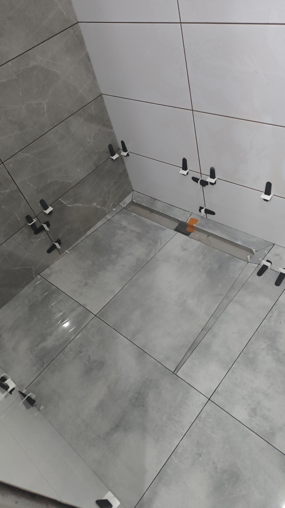 Łazienka w trakcie wykańczania: szare i białe płytki na ścianach i podłodze, z czarnymi klipsami poziomującymi. Widoczny odpływ liniowy. Ukończony etap prac glazurniczych.