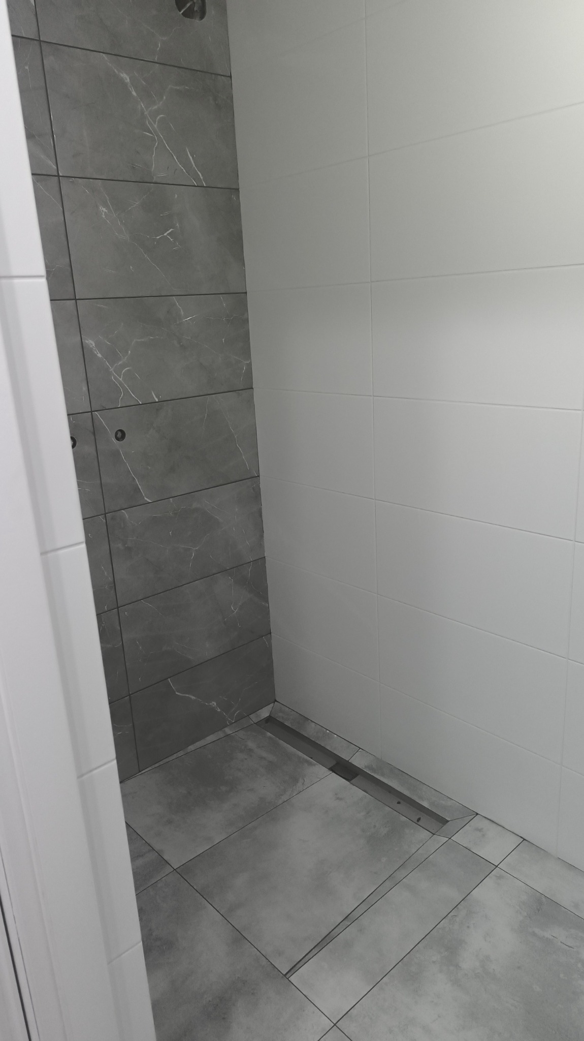 Nowoczesna kabina prysznicowa z szarymi płytkami imitującymi marmur na ścianie i podłodze, oraz białymi płytkami na sąsiedniej ścianie. Widoczny odpływ liniowy.