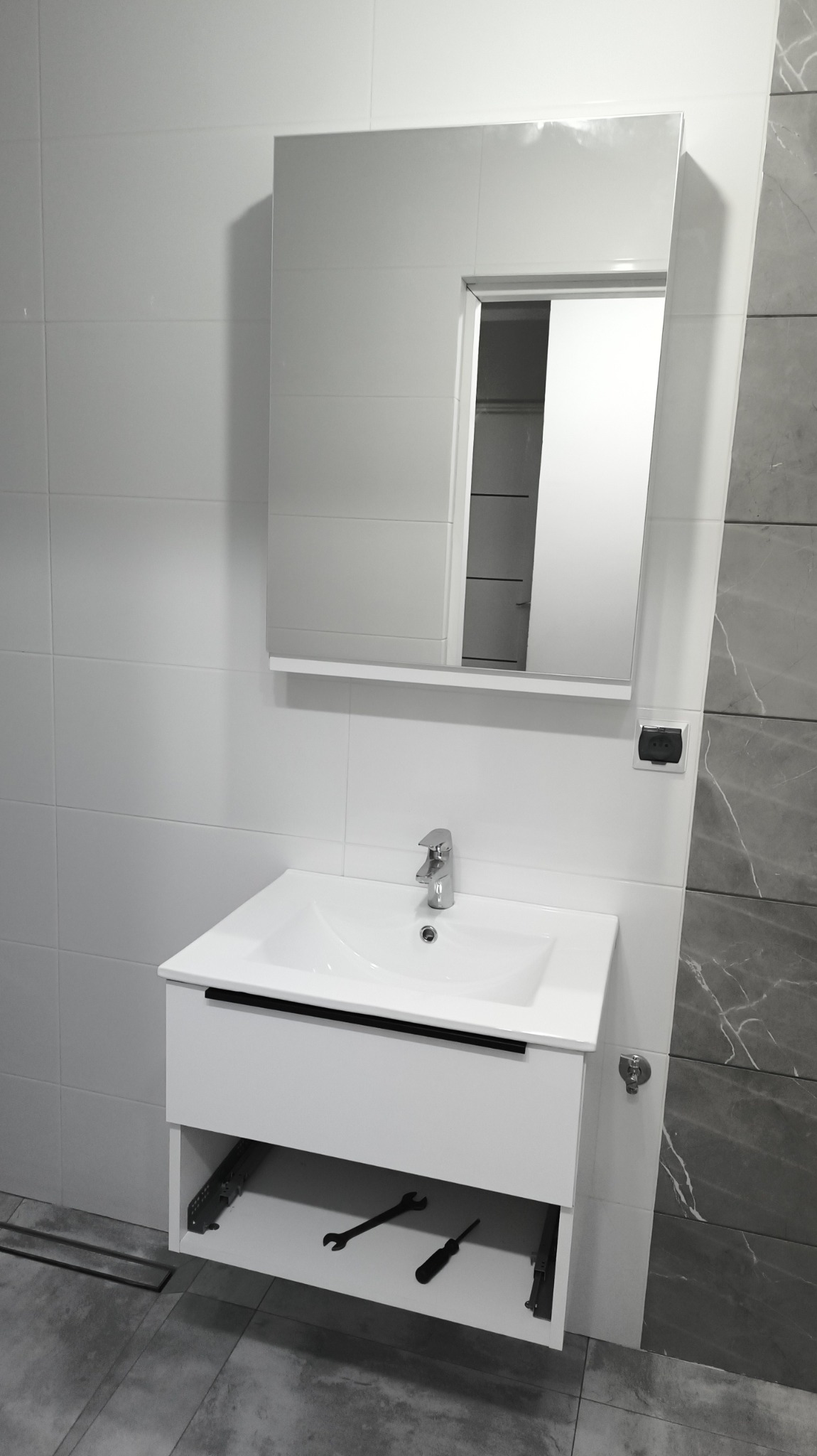 Biała umywalka z szafką i lustrem w nowoczesnej łazience z szarymi płytkami imitującymi marmur. Na półce pod umywalką leżą narzędzia.