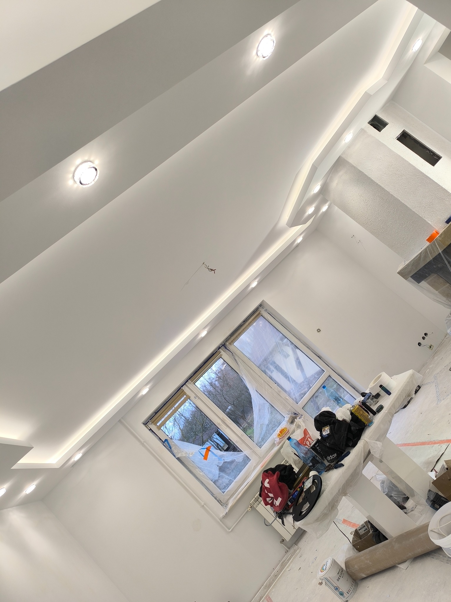 Białe, nowoczesne wnętrze po malowaniu w Długosiodle. Sufit z oświetleniem punktowym i listwami. Okno zabezpieczone folią malarską. Widoczne narzędzia i materiały malarskie.