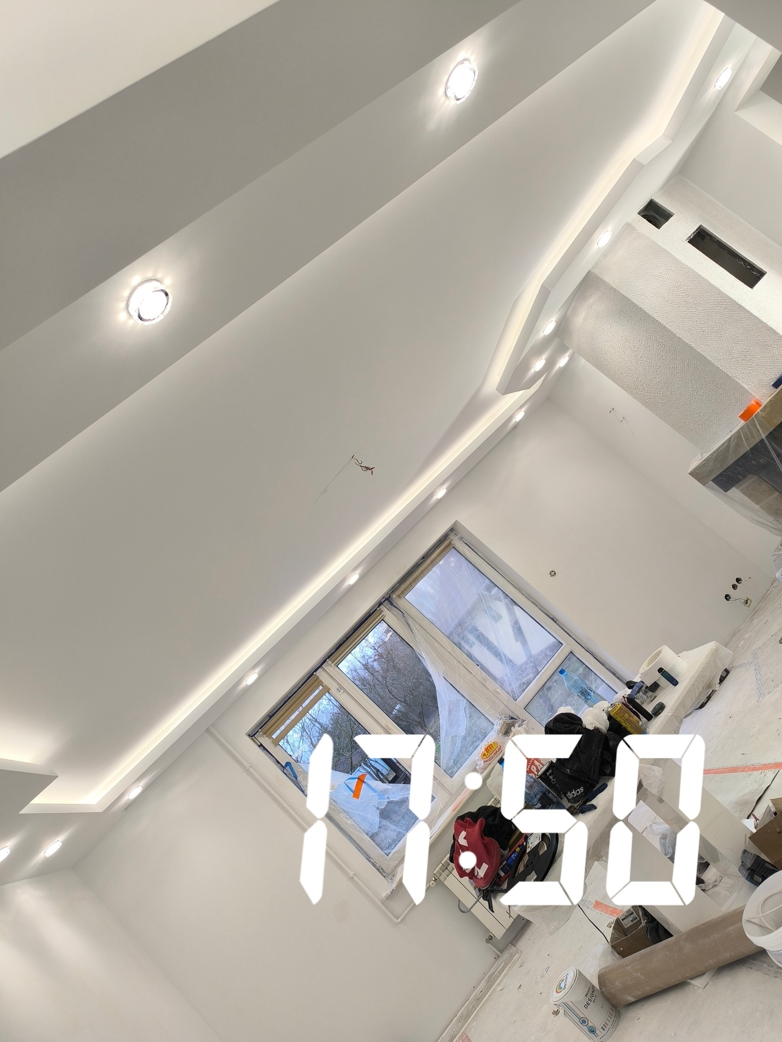 Białe wnętrze po remoncie z oknem, widoczne wykończone ściany, sufit z oświetleniem punktowym i częściowo zakryte podłogi. W tle okno z widokiem na drzewa.