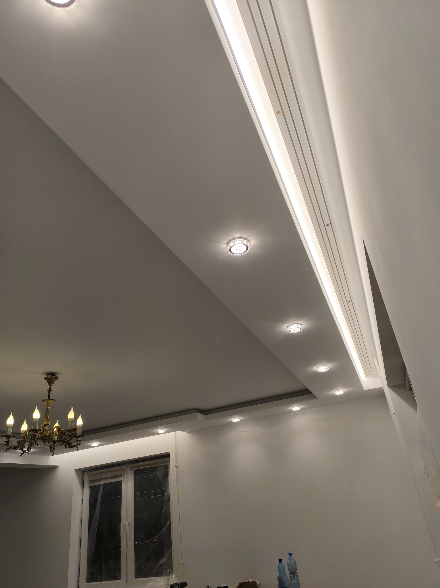 Podwieszany sufit z oświetleniem punktowym i listwą LED, widok z dołu, złoty żyrandol w rogu, okno zabezpieczone folią malarską. Świeżo wykończone wnętrze.