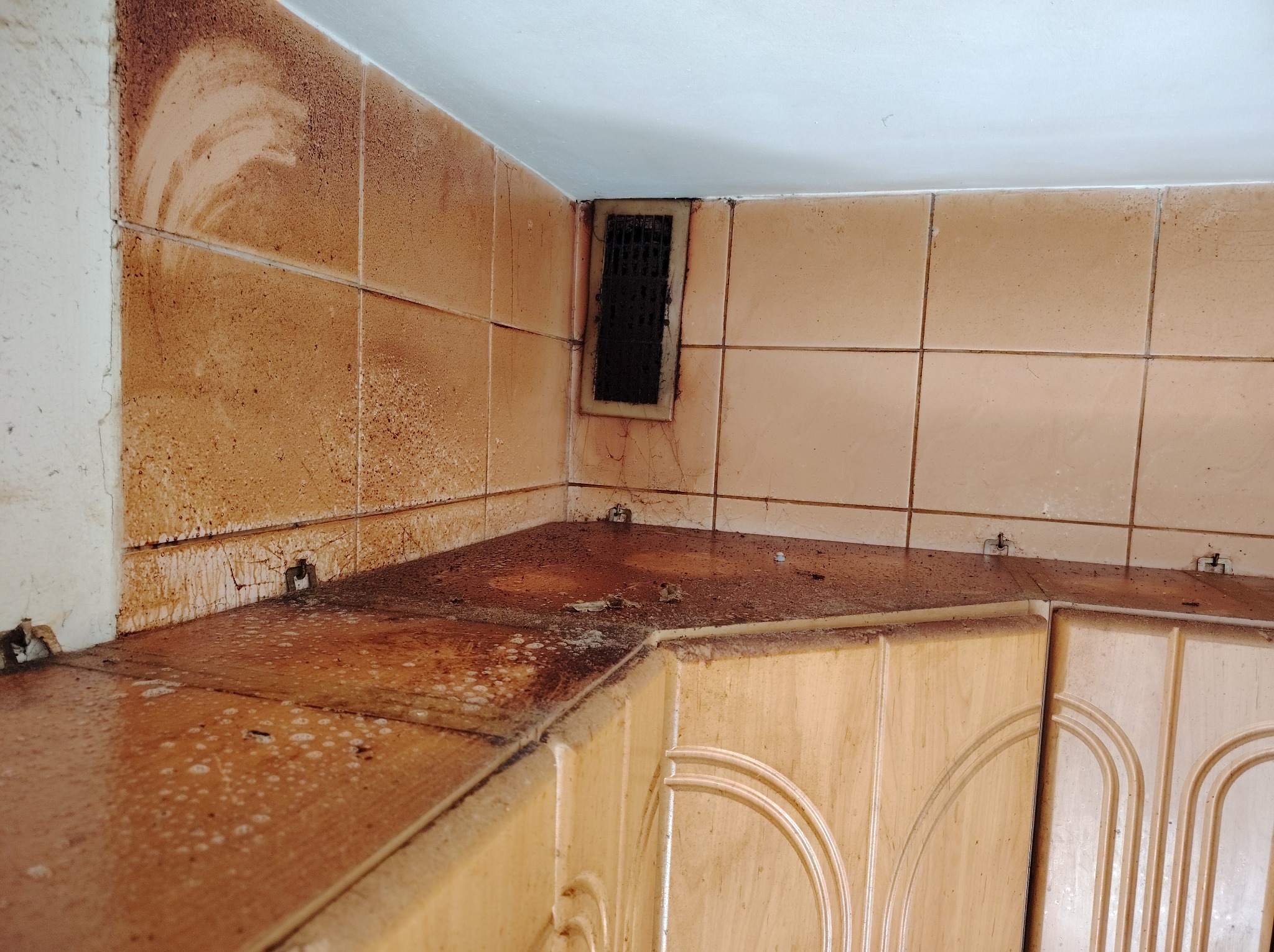 Zabrudzona, zaniedbana kuchnia z brudnymi płytkami, blatem i kratką wentylacyjną. Widoczne ślady tłuszczu i zabrudzeń na powierzchniach. Wymaga gruntownego czyszczenia.