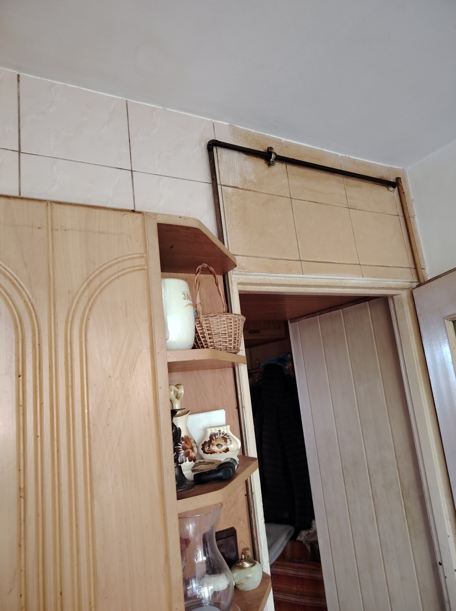 Wnętrze mieszkania z drewnianą szafką, półkami z dekoracjami i otwartymi drzwiami. Widoczne płytki na ścianie i sufit. Ujęcie z dołu, perspektywa z narożnika pokoju.