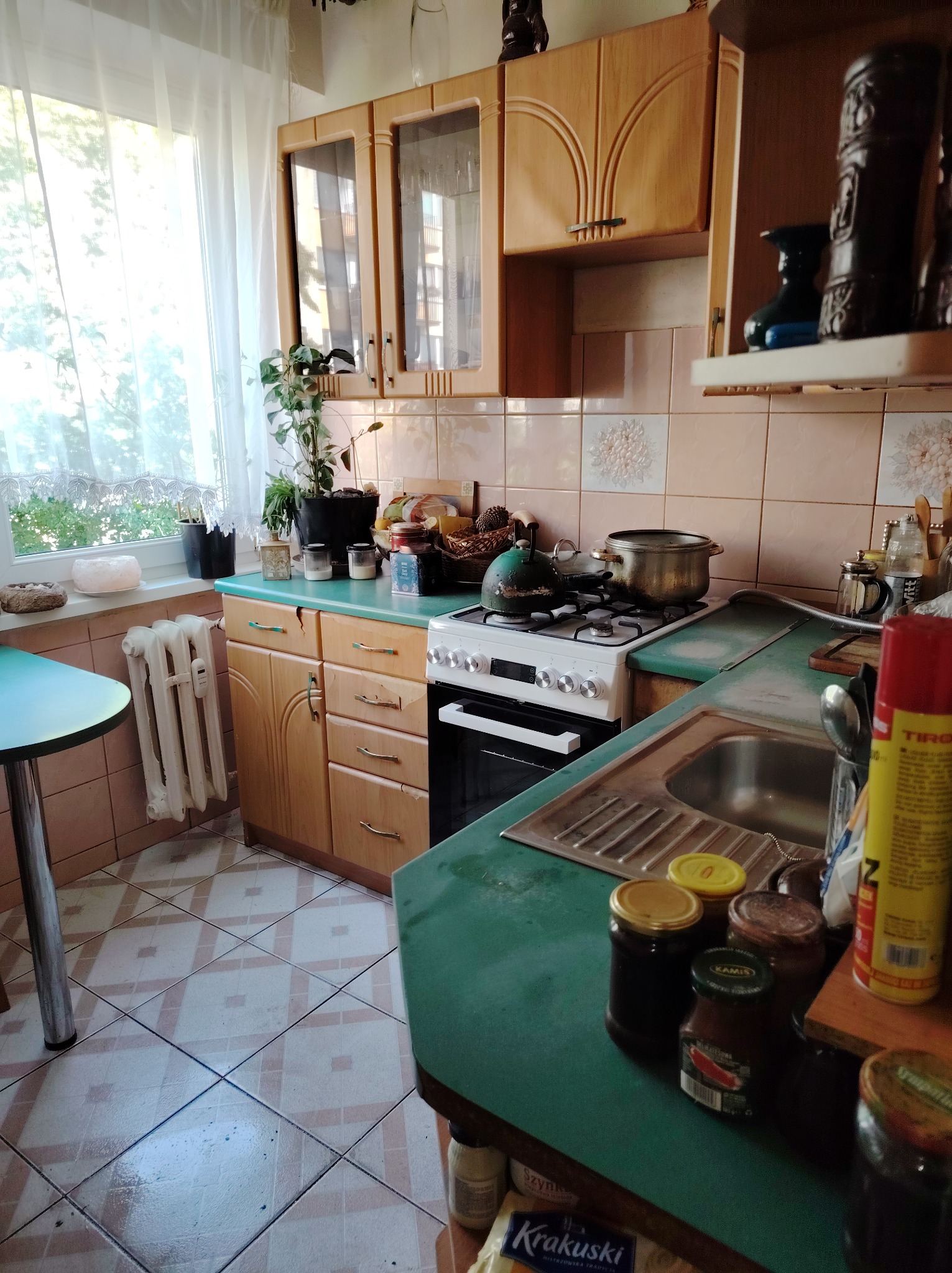 Kuchnia w stylu retro z drewnianymi szafkami, zielonym blatem i wzorzystymi płytkami. Na kuchence stoi garnek, a na blacie słoiki z przetworami. Widoczne okno z firanką.