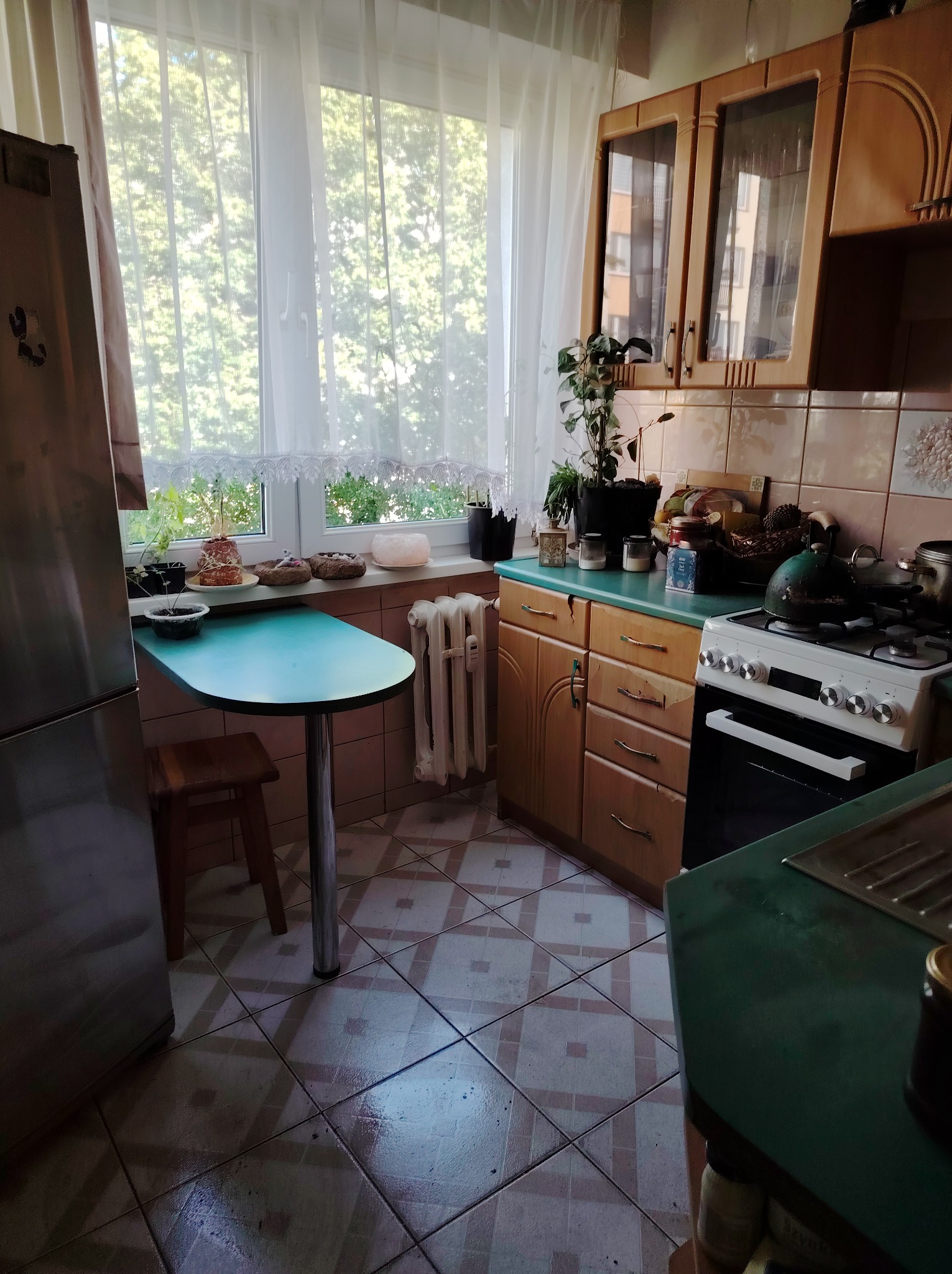 Ustawna kuchnia z oknem, drewnianymi szafkami i turkusowym blatem. Na parapecie rośliny i dekoracje. Na kuchence czajnik. Widoczne płytki podłogowe i grzejnik.