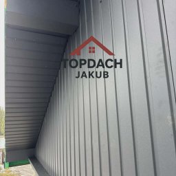 Szara blacha trapezowa na dachu domu, widok z dołu, z widocznym fragmentem elewacji i pracującym dekarzem. Logo firmy nałożone na zdjęcie.