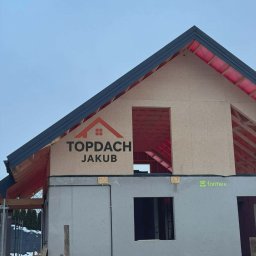 Budowa domu w stanie surowym otwartym z widoczną więźbą dachową i logo firmy Topdach Jakub na ścianie szczytowej. Widoczne elementy konstrukcyjne i izolacja.