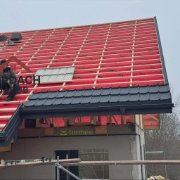 TopDach Jakub Rajczyk - Montaż dachu z czarnej blachy trapezowej. Widoczna konstrukcja drewniana, czerwona membrana i pracownik siedzący na dachu. Częściowo ułożone dachówki.