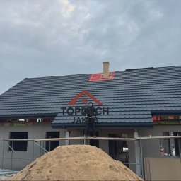 TopDach Jakub Rajczyk - Dom w trakcie budowy z ciemną dachówką, widoczny dekarz na rusztowaniu i sterta piasku. Częściowo wykończony komin z czerwoną izolacją. Pochmurne niebo w tle.