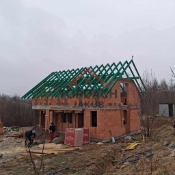 TopDach Jakub Rajczyk - Budowa domu z zieloną więźbą dachową w trakcie montażu. Widoczni dwaj pracownicy, materiały budowlane i otoczenie placu budowy w pochmurny dzień.