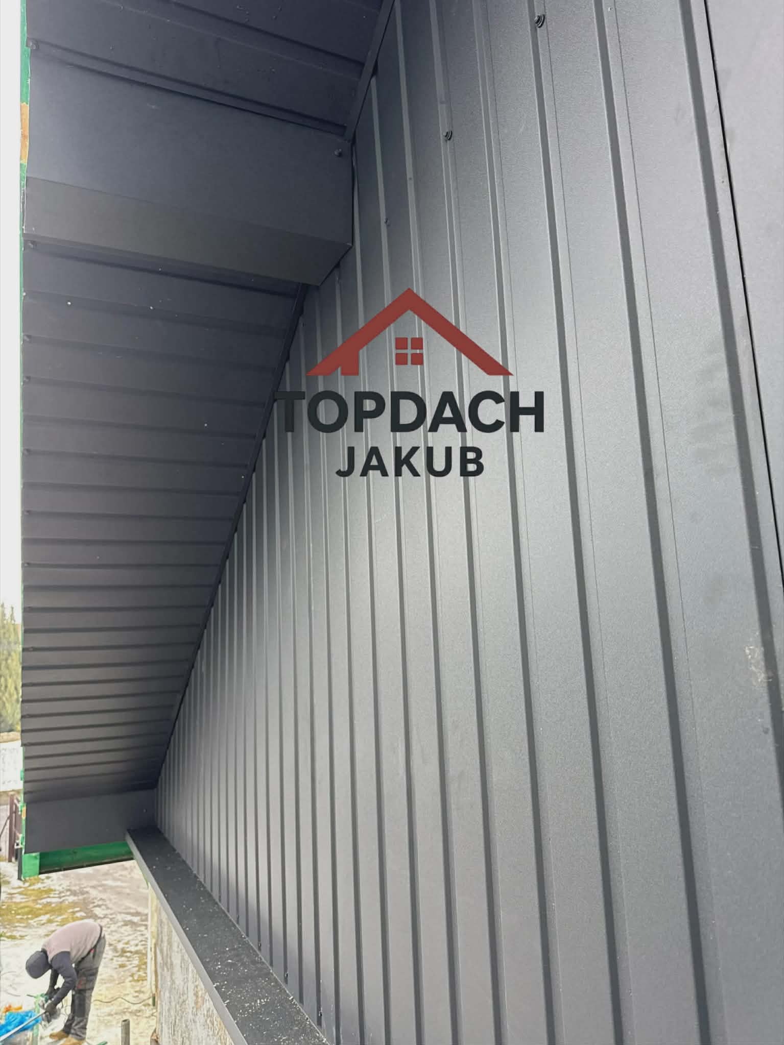 Szara blacha trapezowa na dachu domu, widok z dołu, z widocznym fragmentem elewacji i pracującym dekarzem. Logo firmy nałożone na zdjęcie.