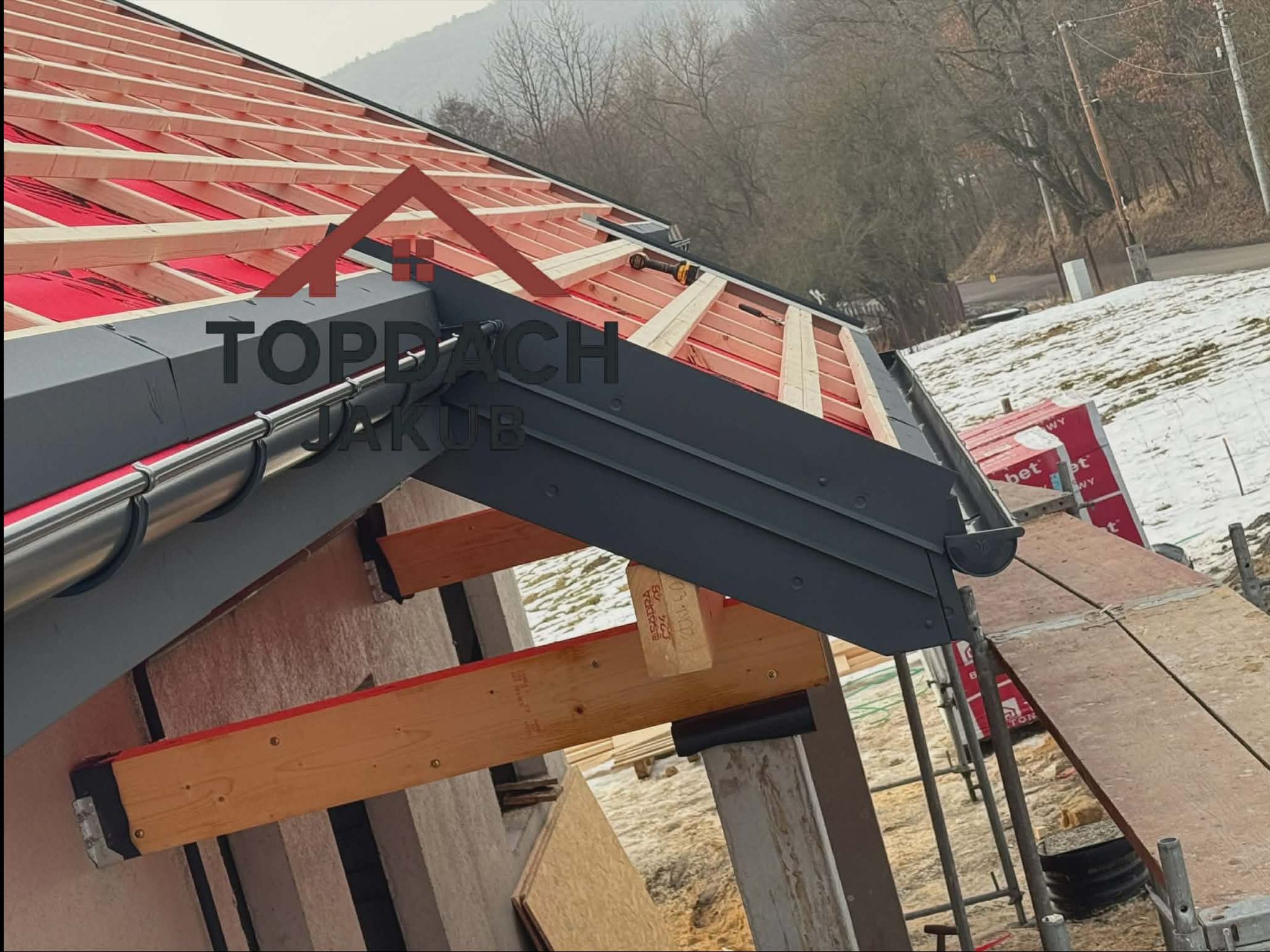 W trakcie budowy dachu: widoczna więźba dachowa z czerwonym pokryciem, szare rynny i elementy konstrukcyjne. W tle zalesione wzgórze i resztki śniegu.