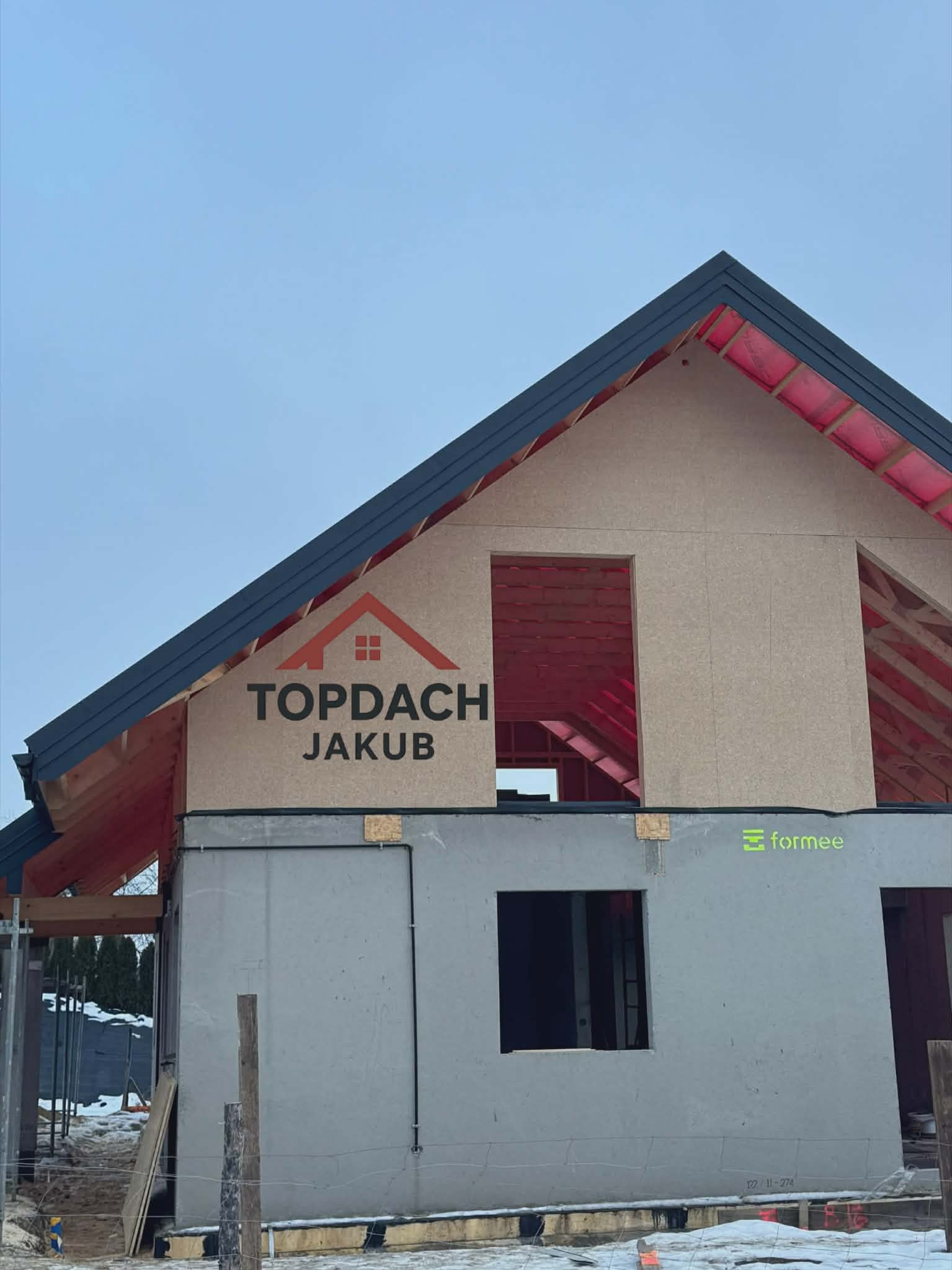 Budowa domu w stanie surowym otwartym z widoczną więźbą dachową i logo firmy Topdach Jakub na ścianie szczytowej. Widoczne elementy konstrukcyjne i izolacja.