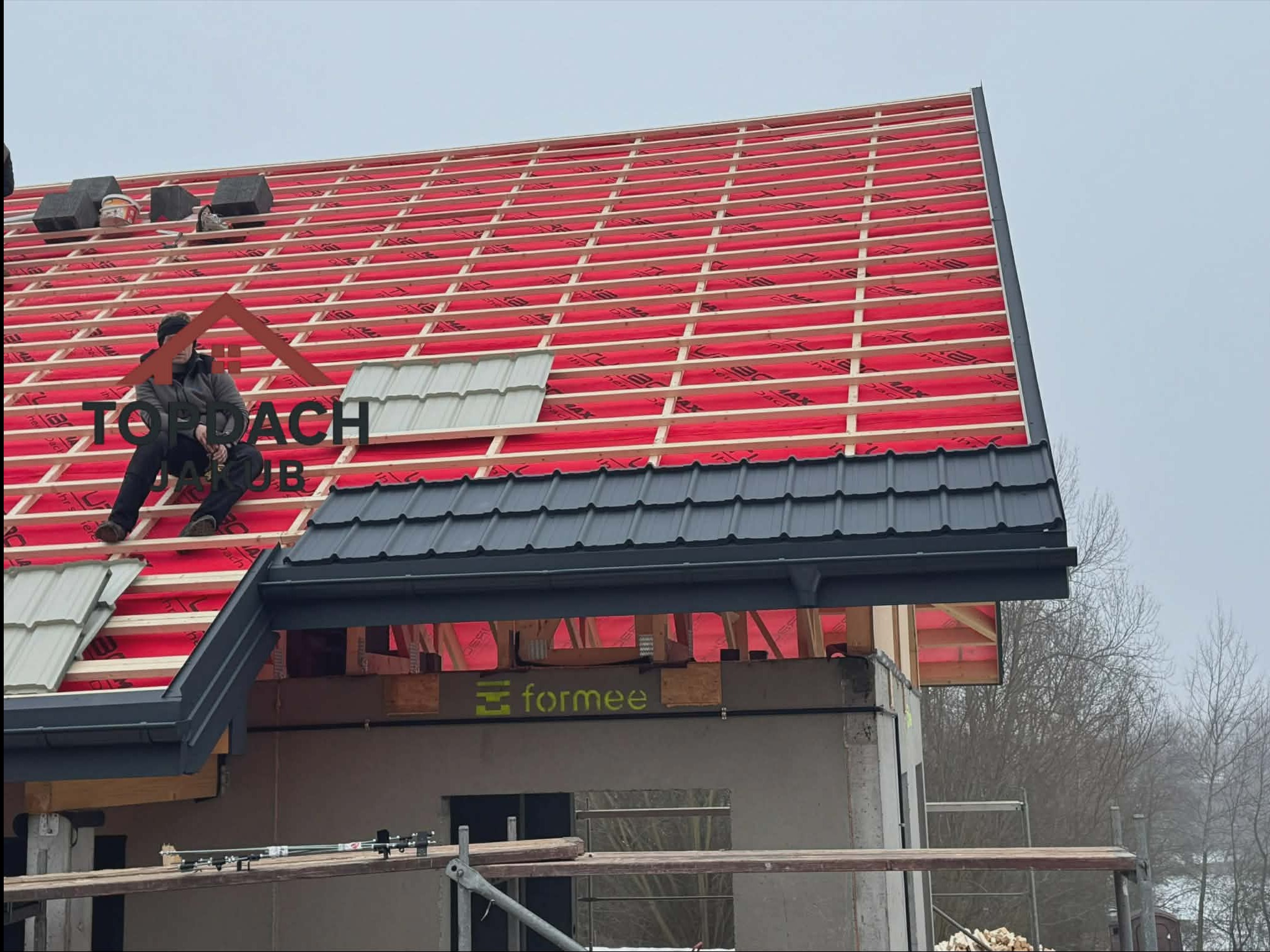 Montaż dachu z czarnej blachy trapezowej. Widoczna konstrukcja drewniana, czerwona membrana i pracownik siedzący na dachu. Częściowo ułożone dachówki.