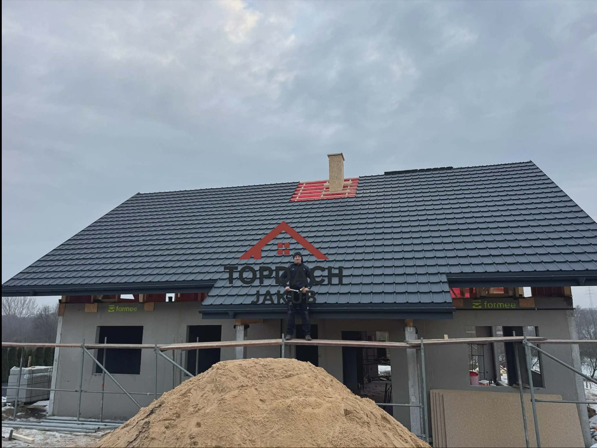 Dom w trakcie budowy z ciemną dachówką, widoczny dekarz na rusztowaniu i sterta piasku. Częściowo wykończony komin z czerwoną izolacją. Pochmurne niebo w tle.