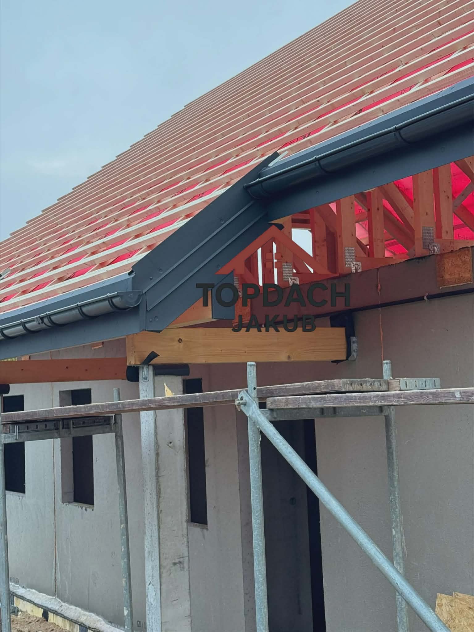 W trakcie budowy: widok na drewnianą konstrukcję dachu z zamontowanymi rynnami w kolorze grafitowym. Widoczne rusztowania i fragment budynku w surowym stanie.