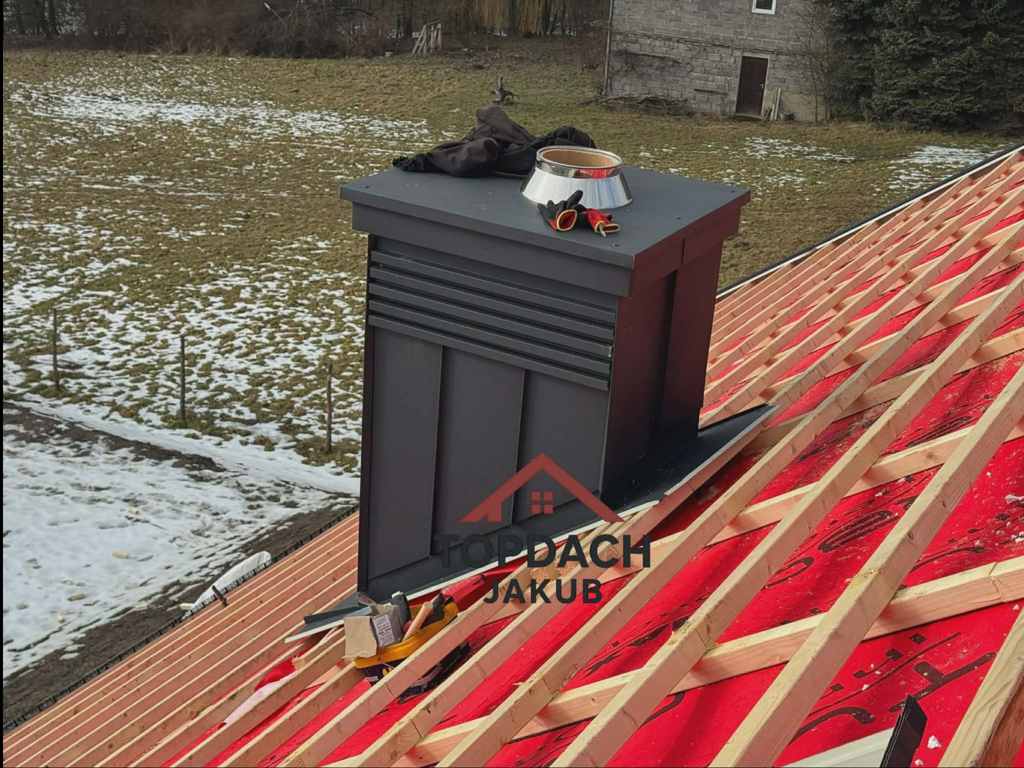 Czarny komin z blachy na dachu w trakcie budowy, widoczna więźba i membrana dachowa. Na kominie leżą rękawice robocze i elementy montażowe. W tle ośnieżone pole i budynek.