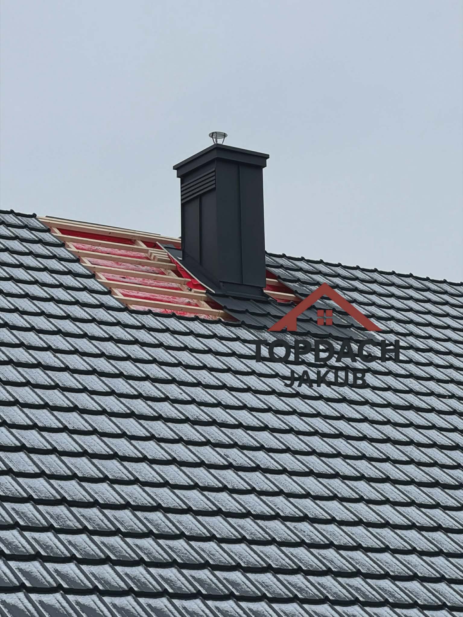 Fragment dachu z czarną blachodachówką, kominem i odkrytą konstrukcją. Widoczna izolacja i łaty. Na dachu logo firmy.