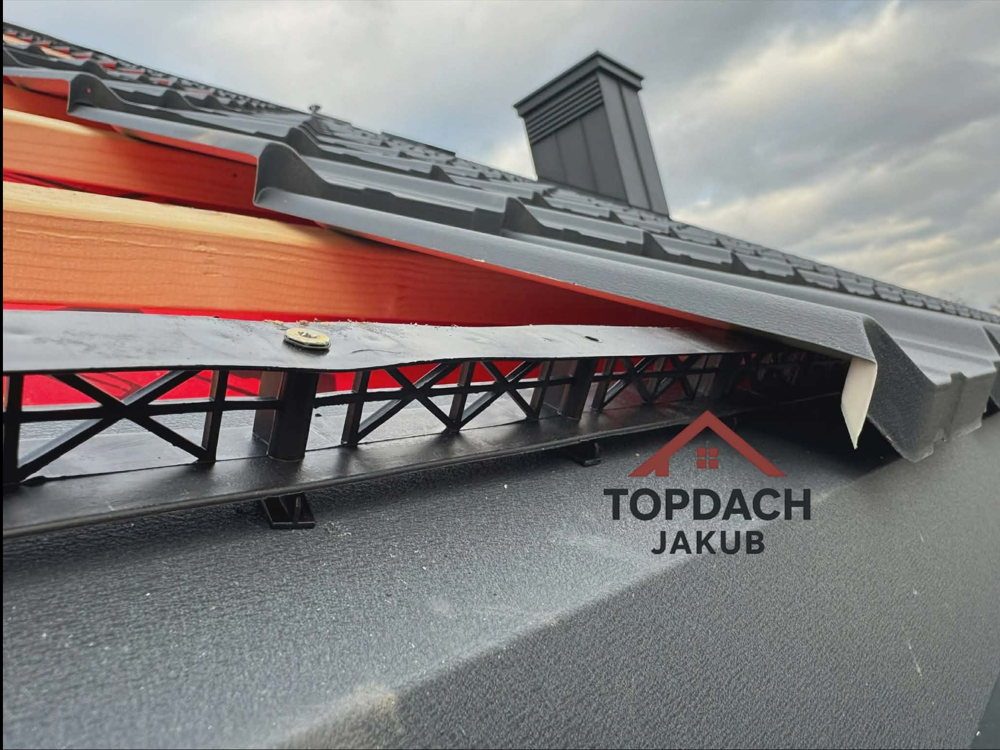 Fragment dachu z czarną blachodachówką, widoczne elementy konstrukcyjne i komin. Ujęcie z dołu, podkreślające detale montażu i wykończenia.