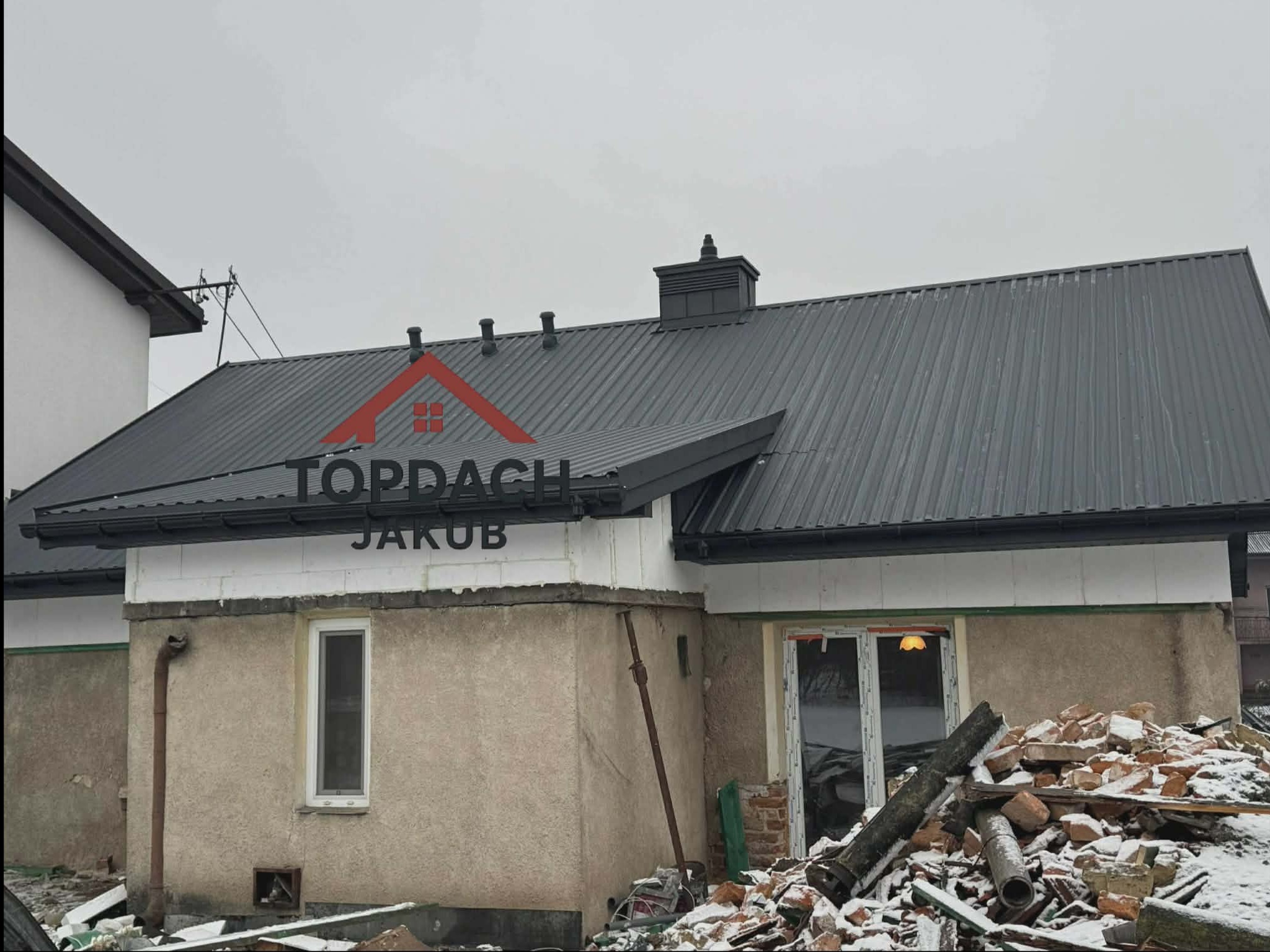 Szary dach z blachy trapezowej po renowacji, widoczny komin i rynny. Na pierwszym planie gruz i śnieg. Logo firmy na elewacji budynku.