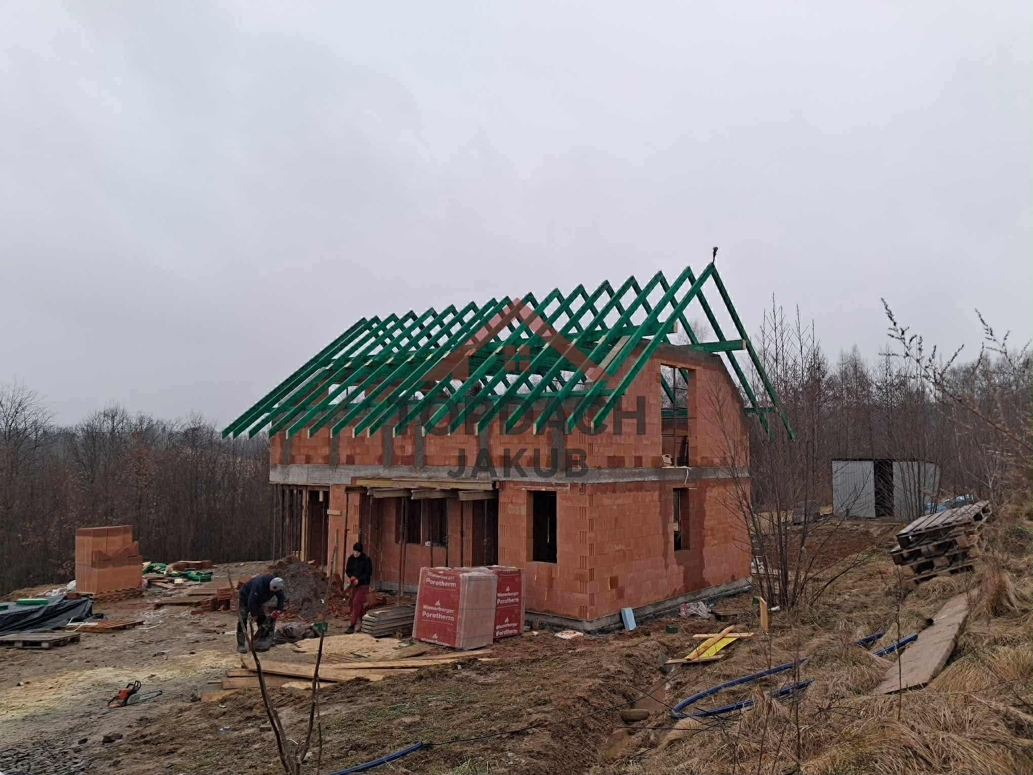 Budowa domu z zieloną więźbą dachową w trakcie montażu. Widoczni dwaj pracownicy, materiały budowlane i otoczenie placu budowy w pochmurny dzień.