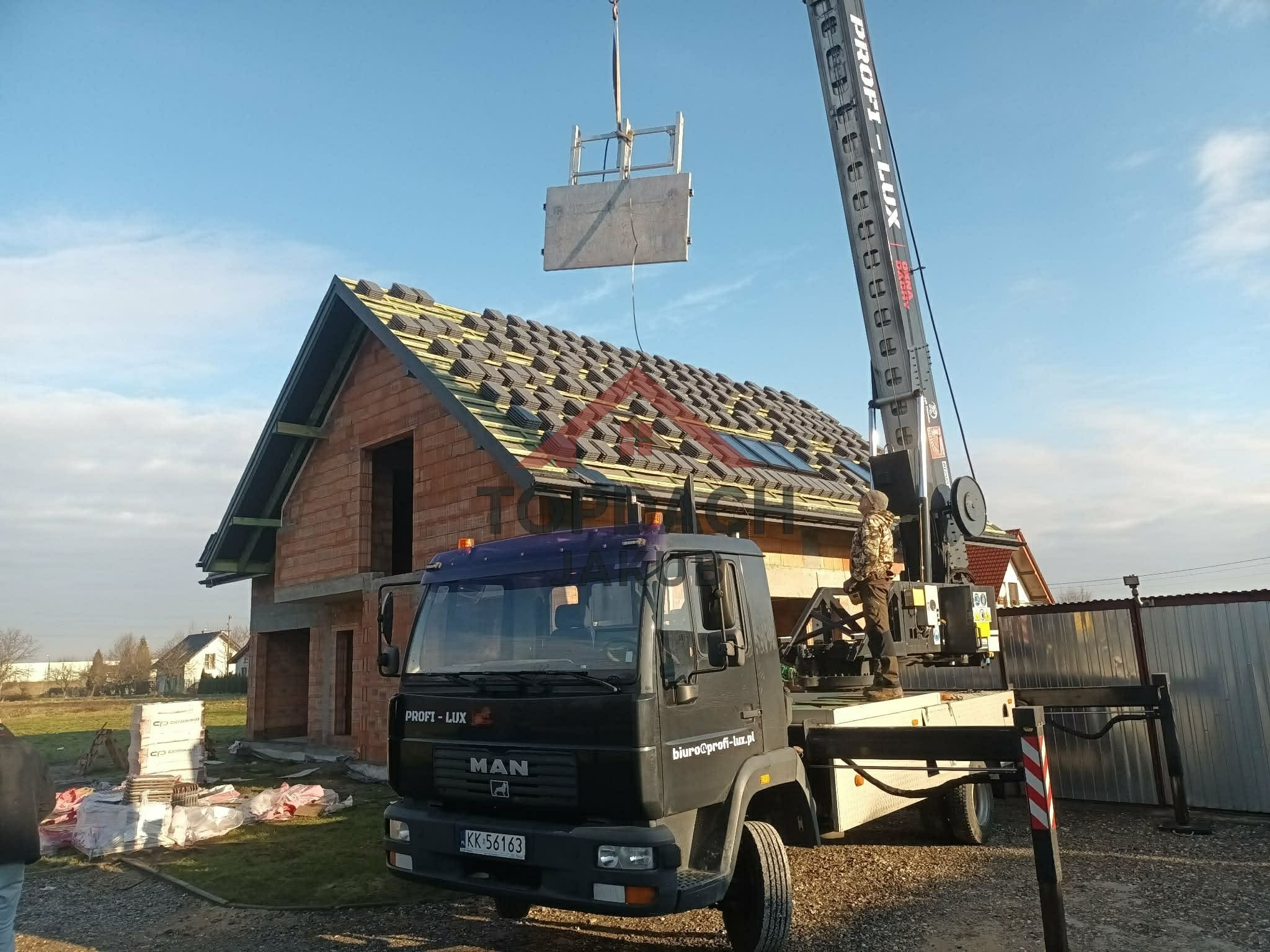 Montaż dachu z użyciem dźwigu na budowie domu jednorodzinnego. Widoczne elementy konstrukcyjne dachu i częściowo ułożona dachówka. Na pierwszym planie ciężarówka z dźwigiem.