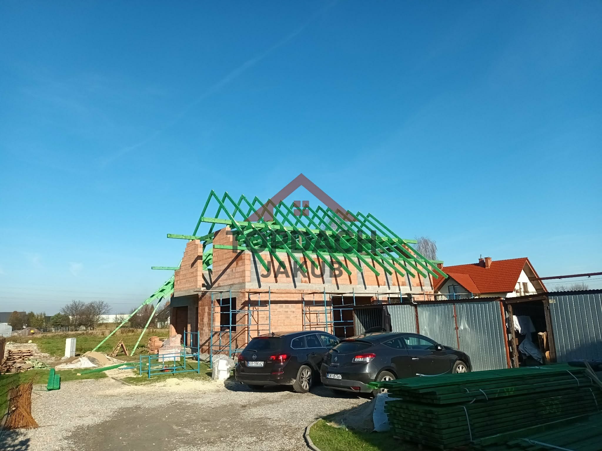 Budowa domu jednorodzinnego w trakcie wznoszenia więźby dachowej z zielonych belek. Na pierwszym planie zaparkowane auta i materiały budowlane.