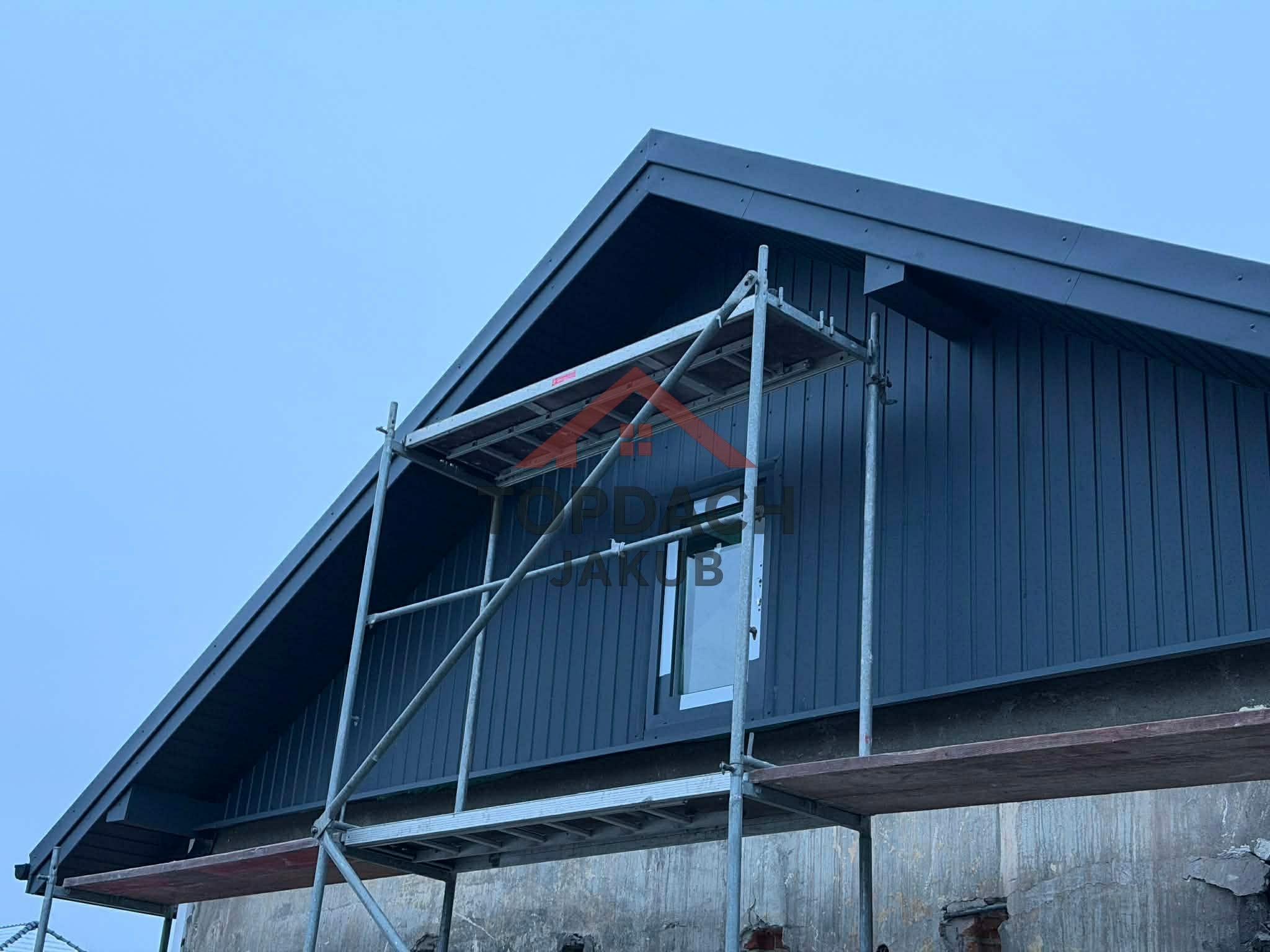 Nowoczesny dom z ciemną elewacją i dachem, widziany z rusztowania. Widoczne detale wykończenia i okno. Niebieskie niebo w tle.