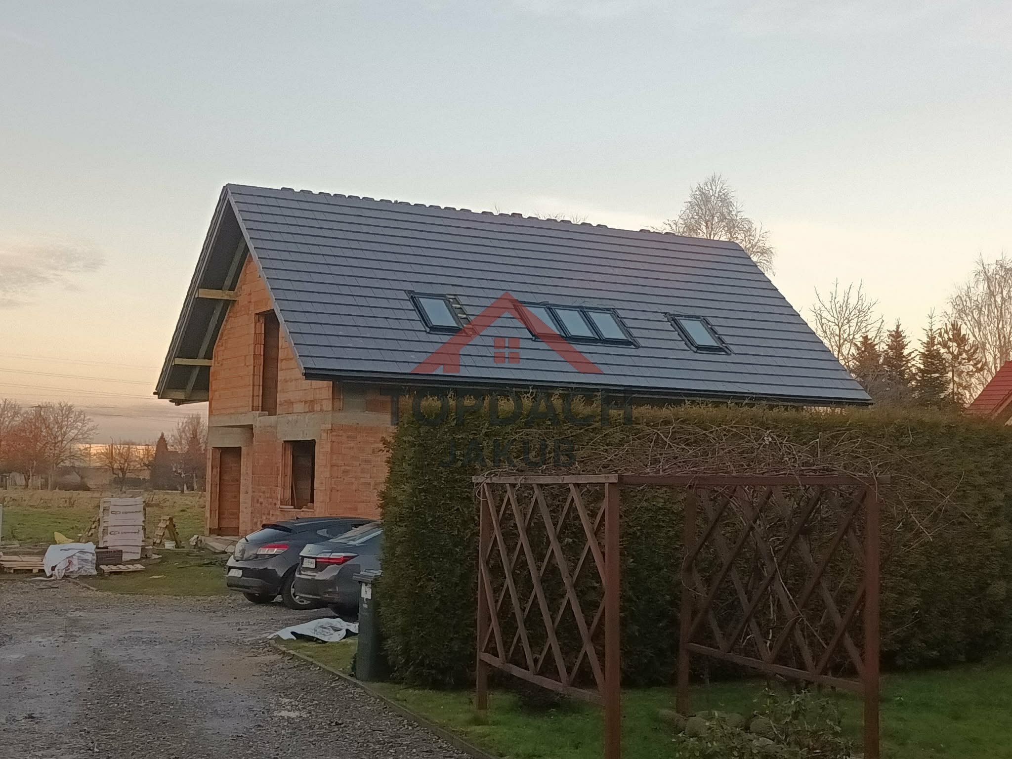 Dom w trakcie budowy z szarym dachem i oknami dachowymi. Widoczne materiały budowlane i dwa samochody. Ujęcie z poziomu gruntu, w tle drzewa i zachmurzone niebo.