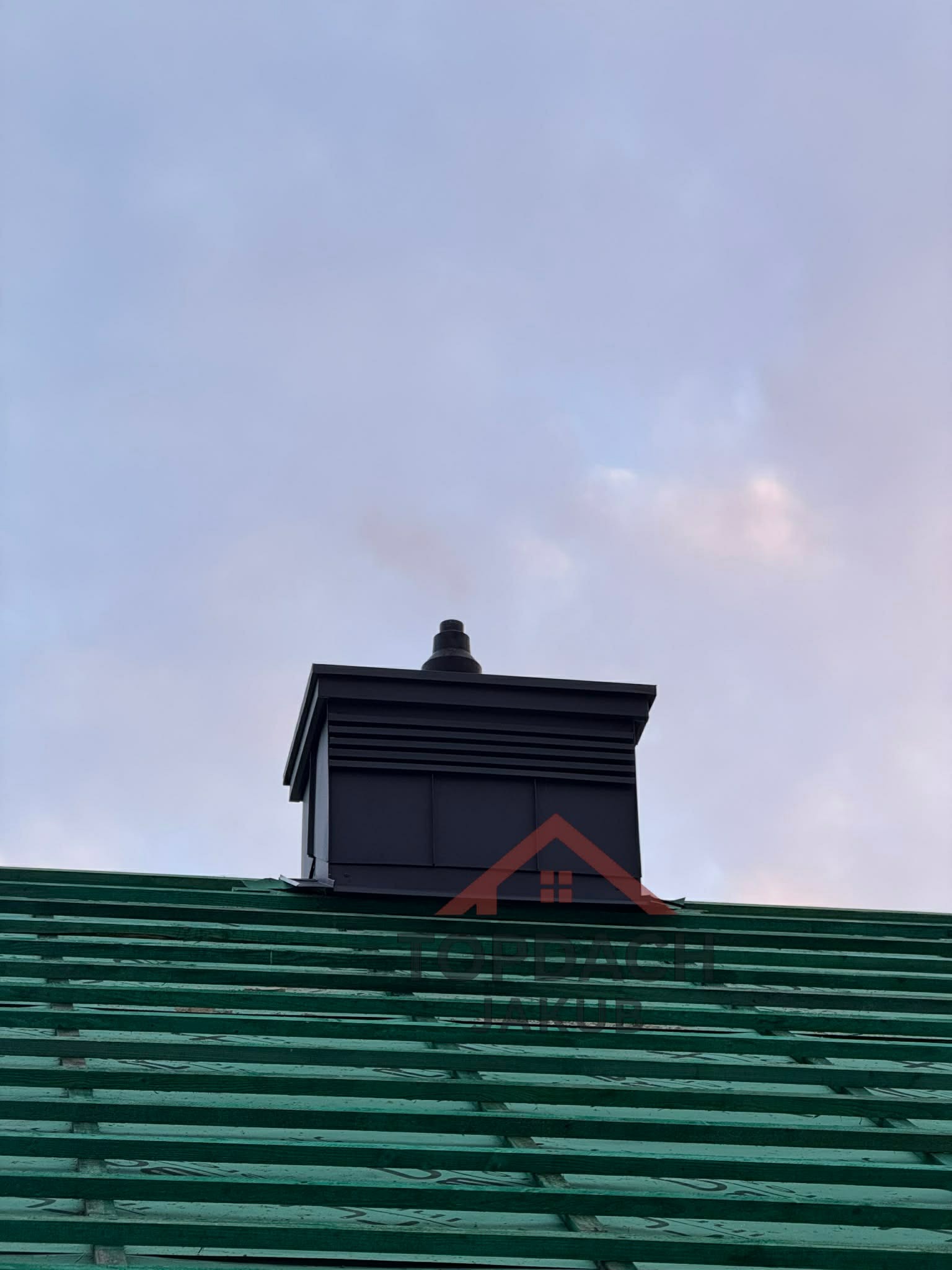 Czarny komin wentylacyjny na zielonym dachu z drewnianą konstrukcją, na tle pochmurnego nieba. Widok z dołu, podkreślający detale konstrukcyjne.
