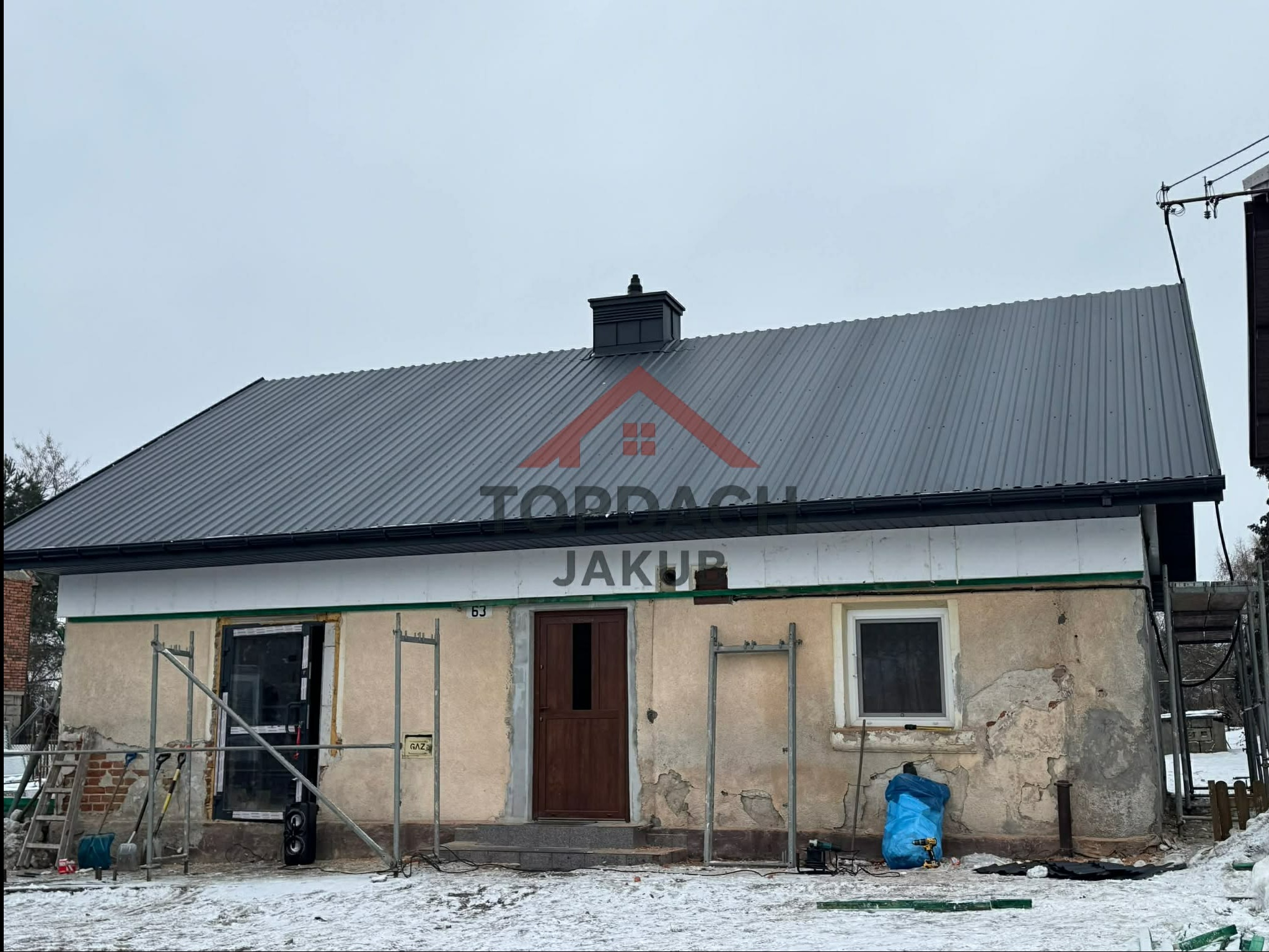 Dom z nowym, szarym dachem z blachy trapezowej w trakcie prac wykończeniowych elewacji. Widoczne rusztowania, narzędzia i ocieplenie. Zimowy krajobraz.