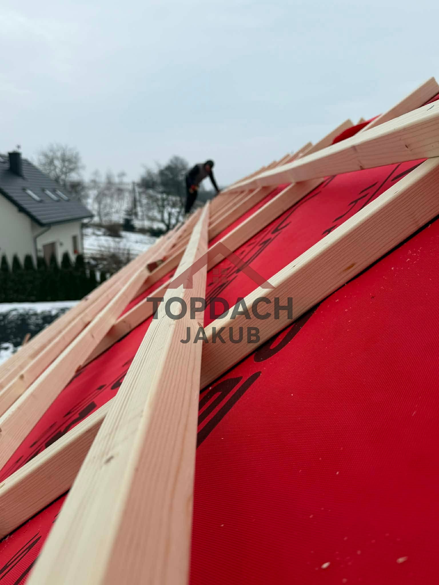 Konstrukcja drewniana dachu pokryta czerwoną membraną, w tle sylwetka pracownika i zabudowa jednorodzinna w pochmurny dzień. Widok z dołu.