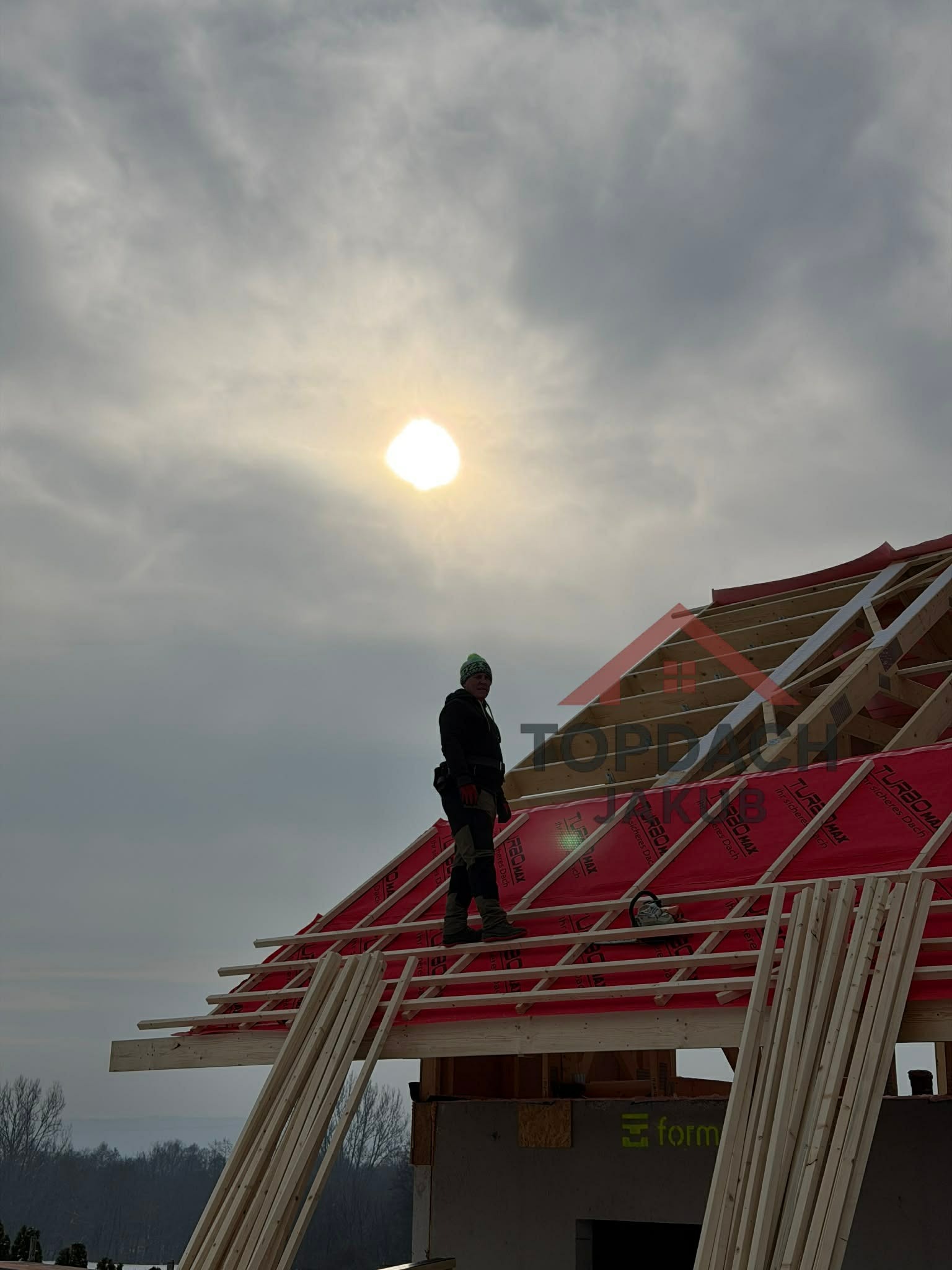 Konstrukcja dachu z czerwoną membraną, na której stoi dekarz w czarnym ubraniu. W tle pochmurne niebo i słońce. Widoczne drewniane elementy więźby i narzędzia.