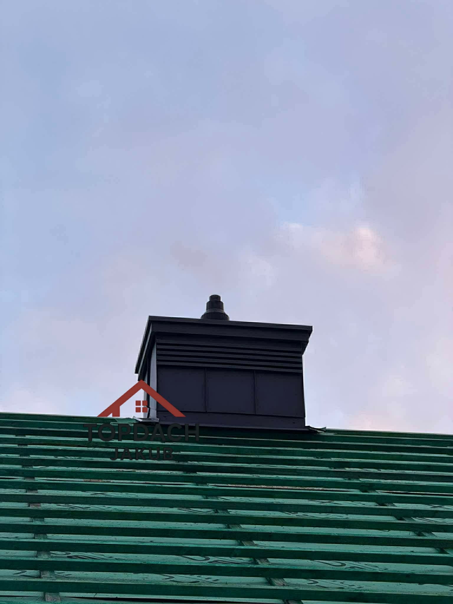 Nowy, czarny komin wentylacyjny na zielonym dachu z łatami, widziany pod kątem. Komin o prostej, nowoczesnej formie, na tle pochmurnego nieba.