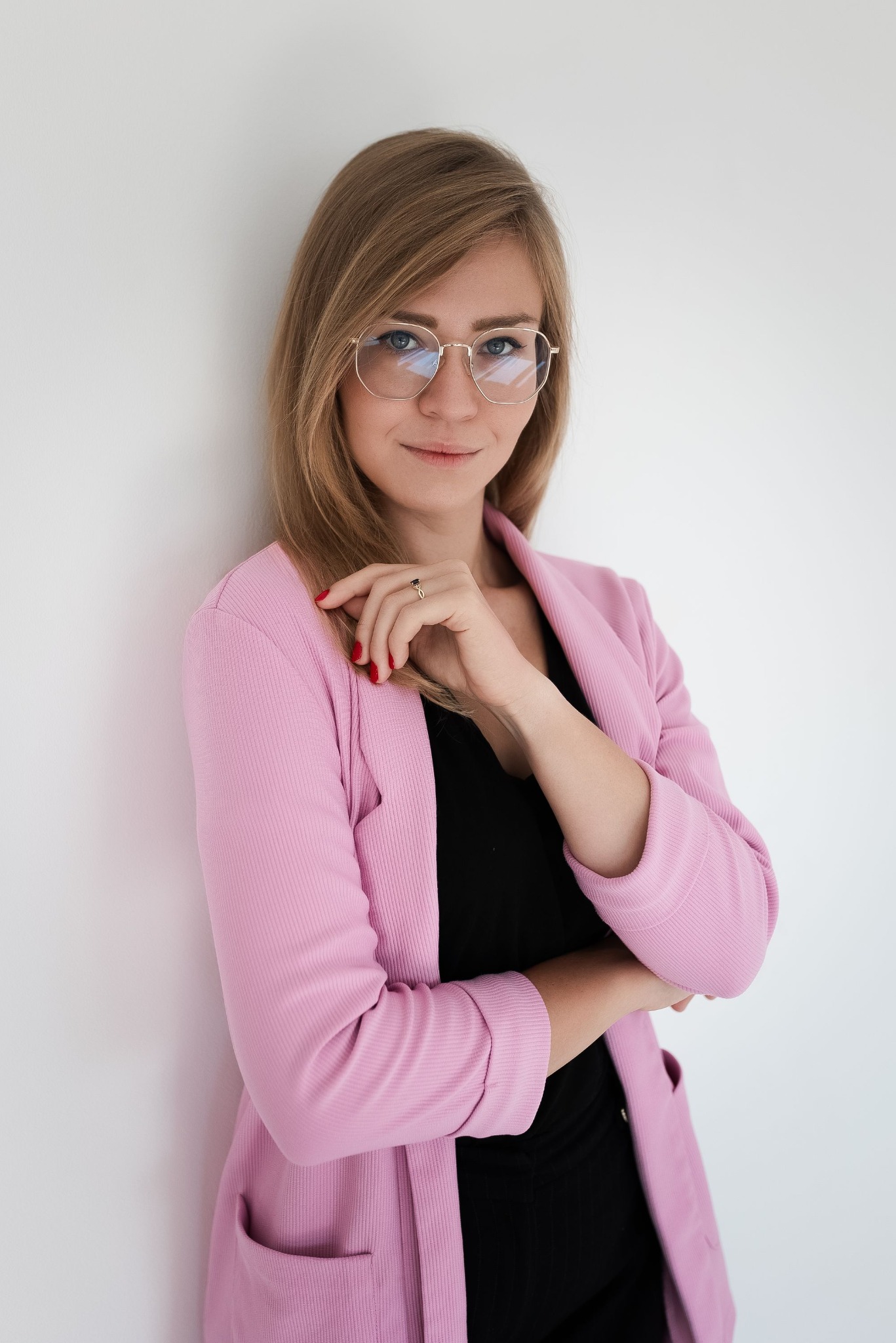 Portret kobiety w okularach i różowym żakiecie, opierającej się o białą ścianę. Uśmiech, naturalne światło, profesjonalny wygląd.