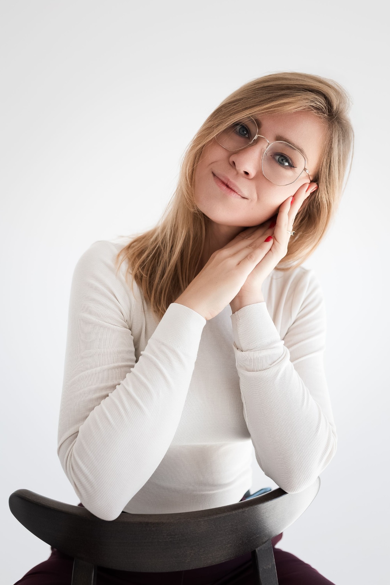 Portret uśmiechniętej kobiety w okularach, siedzącej na krześle, z dłońmi przy twarzy. Jasne tło, naturalne światło, subtelny makijaż, sweter z długim rękawem.