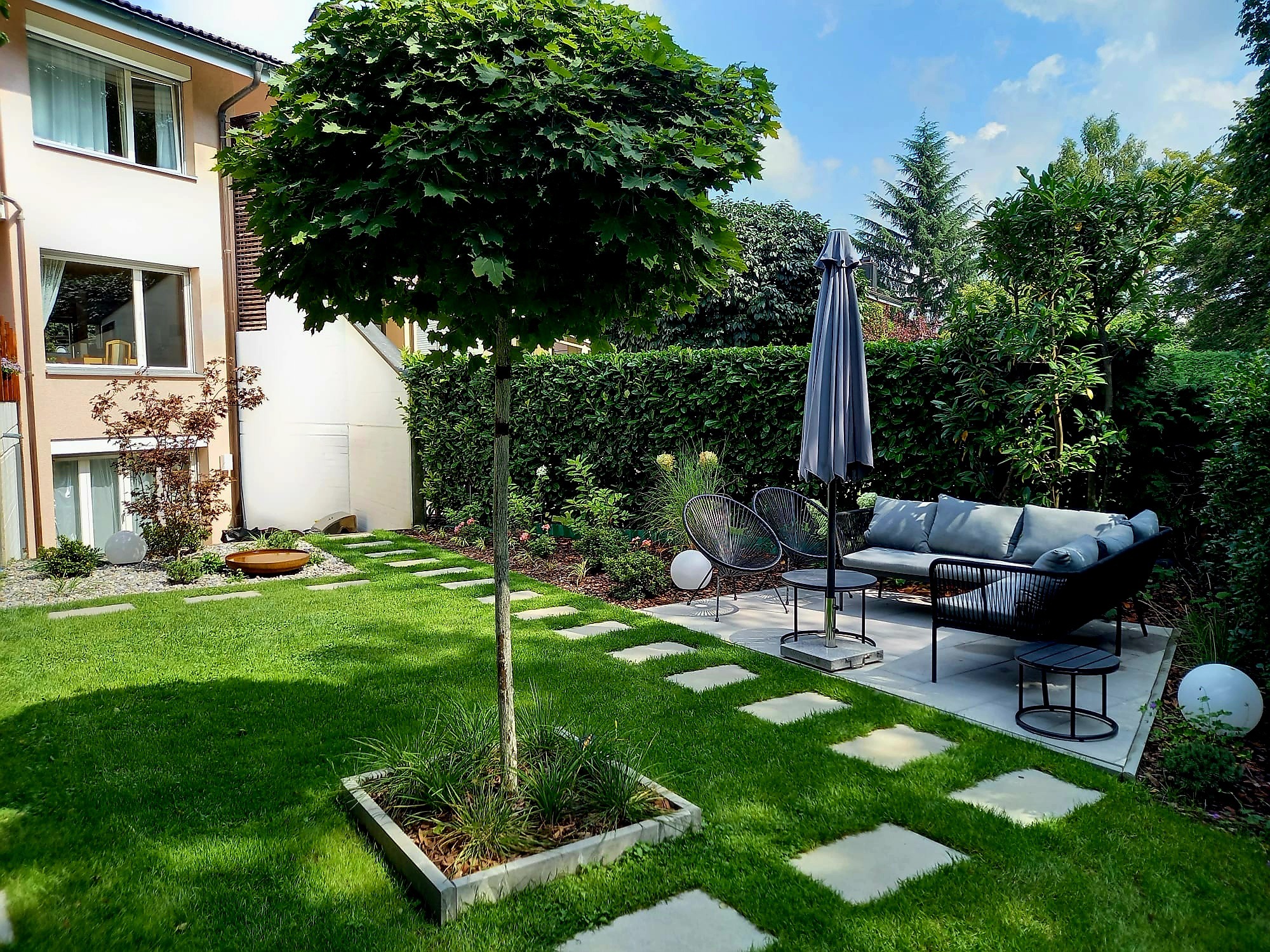 Nowoczesny ogród z równo przystrzyżonym trawnikiem, ścieżką z płyt, drzewem w kwadratowej donicy i meblami ogrodowymi na betonowym patio.