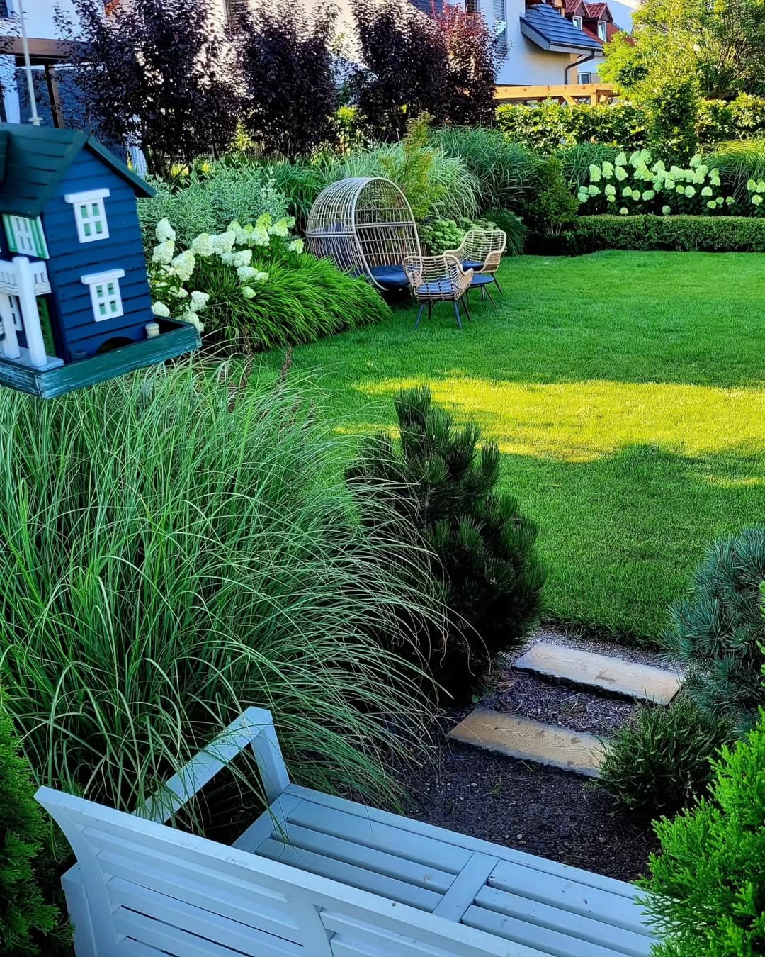 Zadbany ogród z zielonym trawnikiem, ozdobnymi krzewami, ścieżką z kamieni i wiklinowym fotelem wiszącym. W tle widoczny domek dla ptaków i fragment budynku.