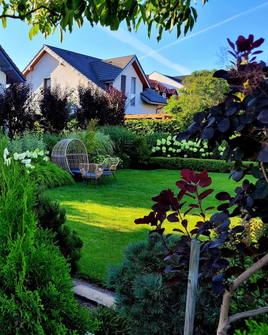 Zadbany ogród z zielonym trawnikiem, ozdobnymi krzewami i kwiatami. W tle dom z ciemnym dachem. Na trawniku okrągłe, wiklinowe fotele z poduszkami.