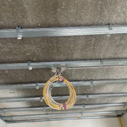 ELMARB Marcin Baran - Kable elektryczne zwisające z sufitu podwieszanego z metalową konstrukcją. Widok z dołu na szare, surowe wykończenie i żółte przewody. Etap prac instalacyjnych.