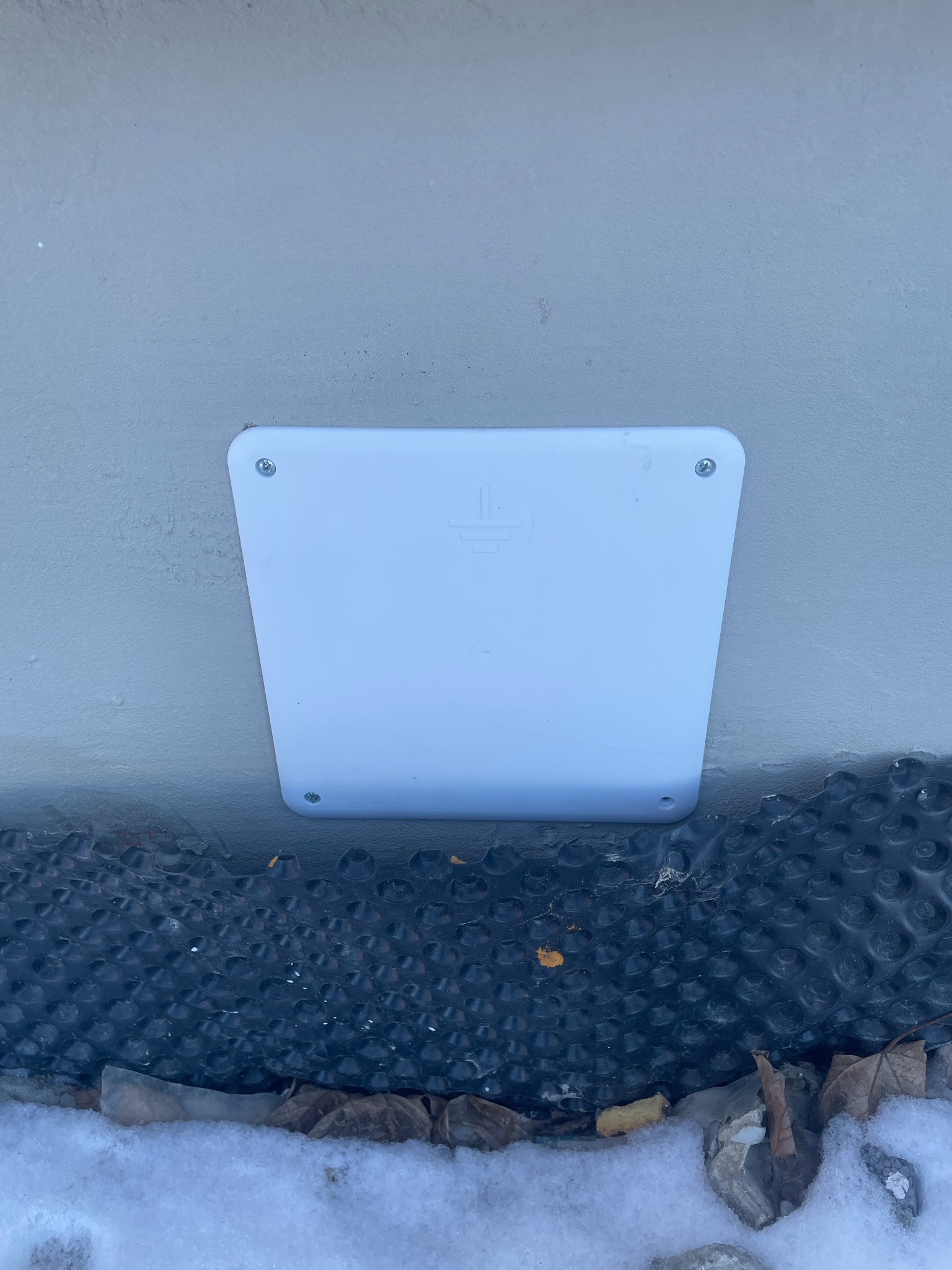 Biała puszka wyrównania potencjałów na szarej ścianie, przymocowana czterema śrubami, z symbolem uziemienia. U dołu folia kubełkowa i śnieg.