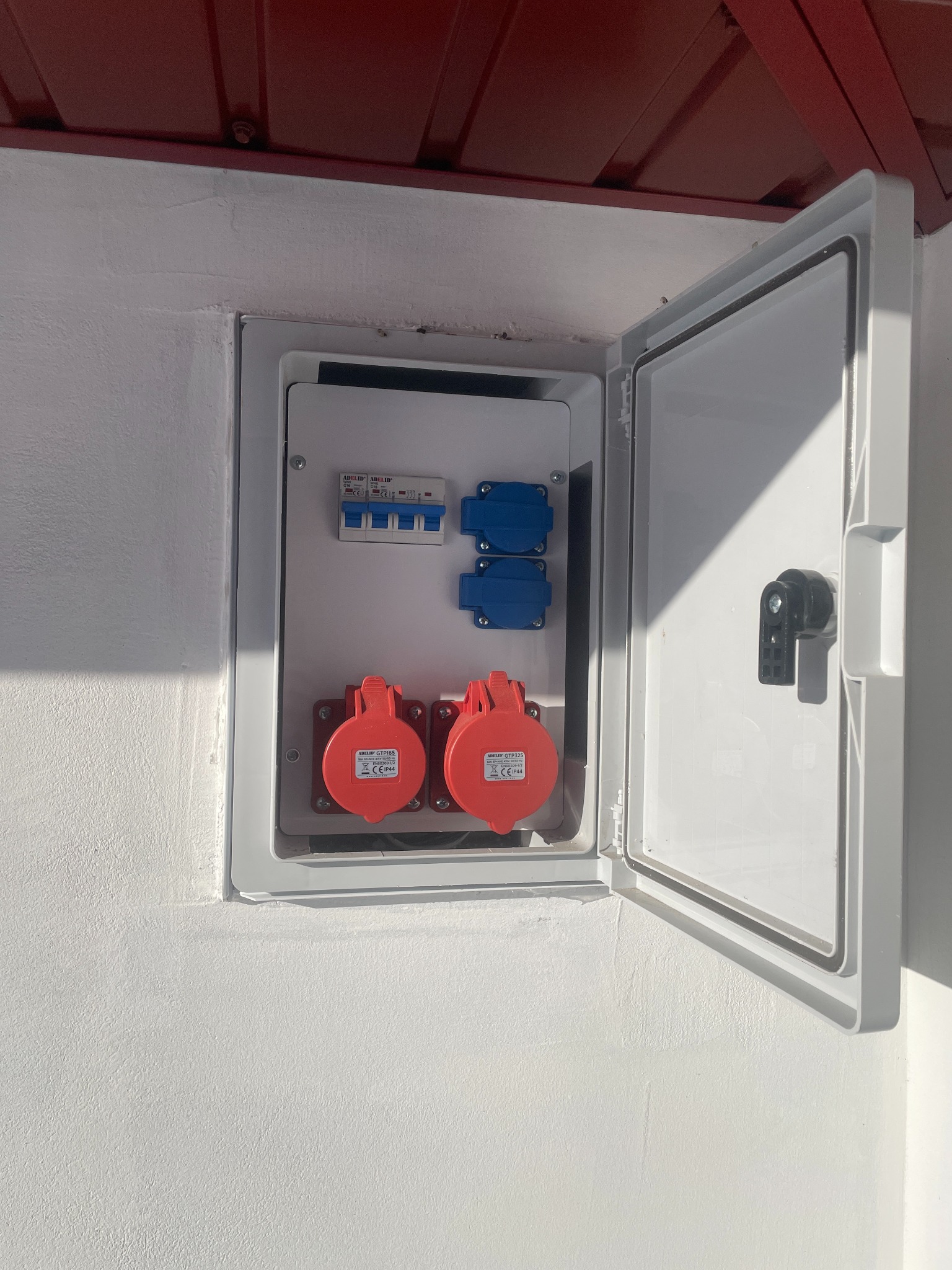 Otwarta skrzynka elektryczna z bezpiecznikami i gniazdami siłowymi (czerwone i niebieskie) na białej ścianie pod czerwonym dachem. Widoczne detale montażu i okablowania.